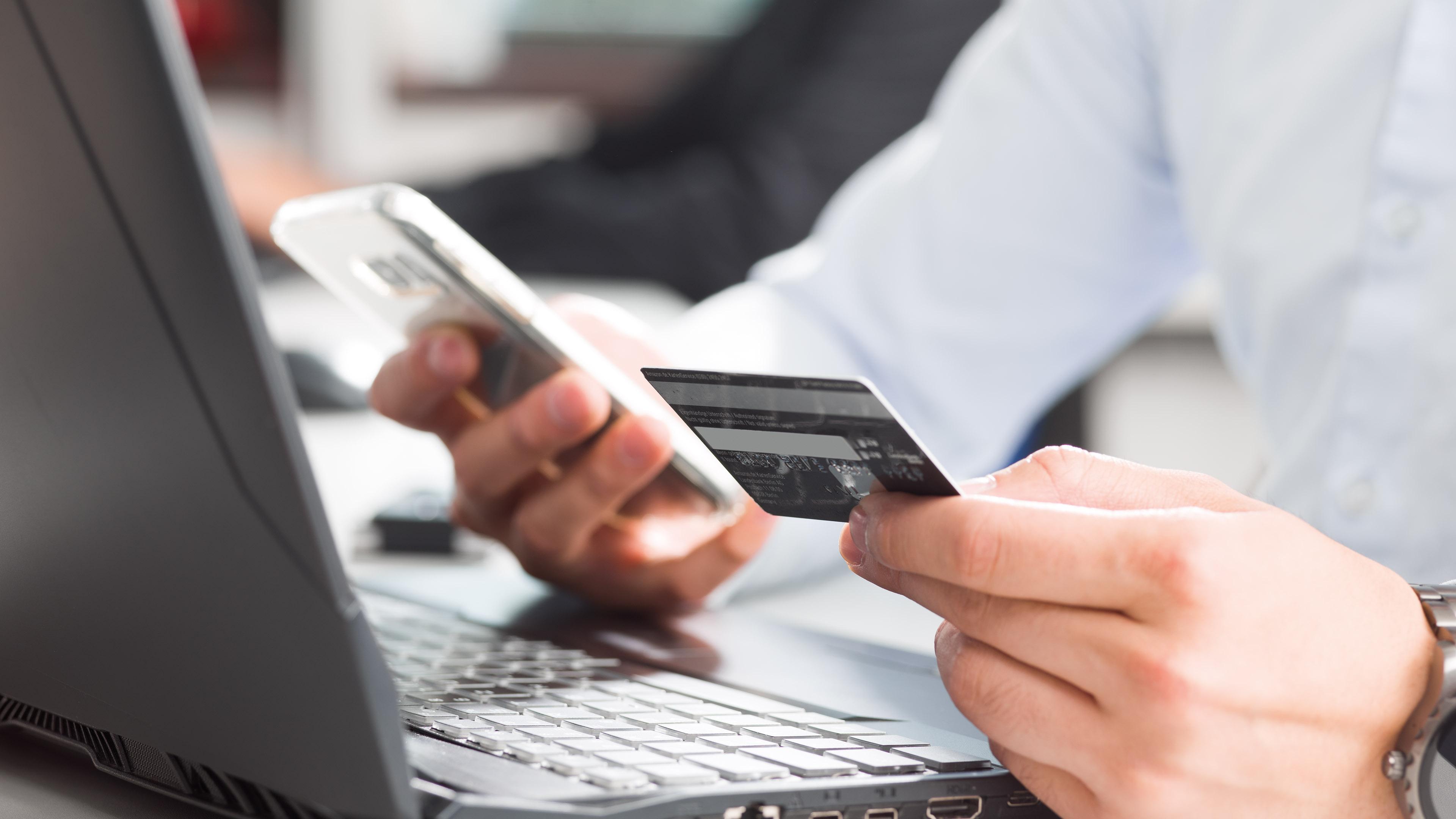 Person hält Smartphone und Kreditkarte vor einem Laptop, bereit für eine Online-Transaktion.