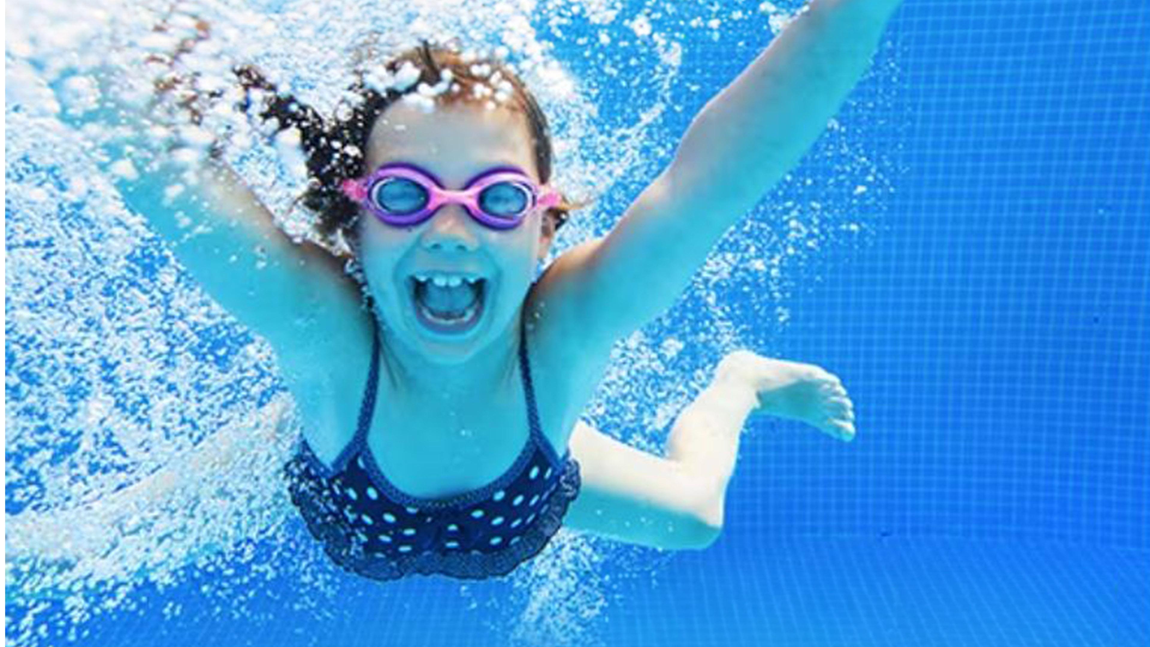 Fröhliches Mädchen mit rosa Schwimmbrille taucht im Pool, spritzendes Wasser, klare blaue Fliesen im Hintergrund.