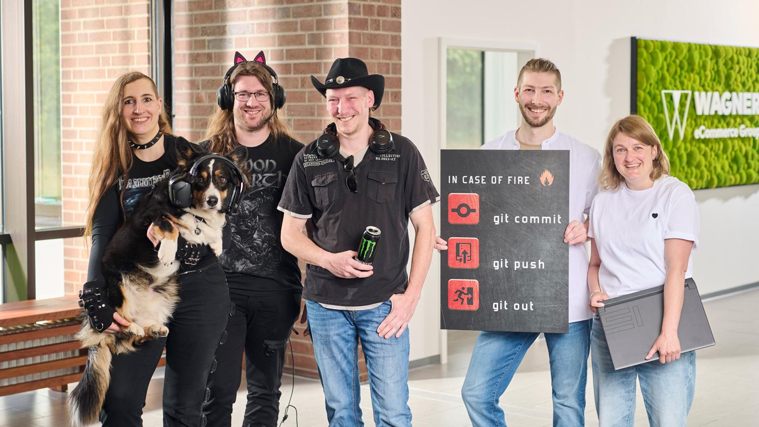 Das Bild zeigt sechs Personen, die in einem modernen Bürogebäude vor einer Wand mit ei em grünen Schild stehen, auf dem 'Wagner eCommerce Group' zu lesen ist. Links steht eine Frau mit langen braunen Haaren, die ein schwarzes Metal-T-Shirt trägt und einen Hund hält, der Kopfhörer trägt. Neben ihr steht ein Mann mit langen Haaren und einer Katzenohren-Kopfbedeckung, der ebenfalls ein schwarzes T-Shirt trägt. Daneben steht ein Mann mit Cowboyhut und Kopfhörern um den Hals, der eine Dose Monster-Energy in der Hand hält. Rechts von ihm steht ein Mann, der ein Schild mit der Aufschrift 'In Case of Fire: git commit, git push, git out' hält. Ganz rechts steht eine Frau mit blonden Haaren und weißem T-Shirt, die ein Laptop unter dem Arm hält. Alle Personen lächeln in die Kamera.
