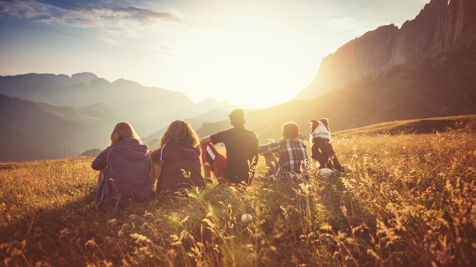 Gruppe von Menschen und ein Hund sitzen auf einer Wiese und genießen den Sonnenuntergang mit Blick auf die Berge.