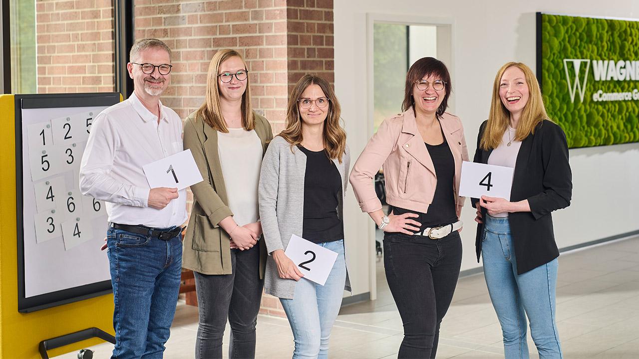 Ein Team von fünf Personen posiert mit nummerierten Karten in einem hellen Büro. Ein Zahlenbrett und ein grünes Wandschild mit der Aufschrift 'Wagner eCommerce Group' sind im Hintergrund sichtbar.