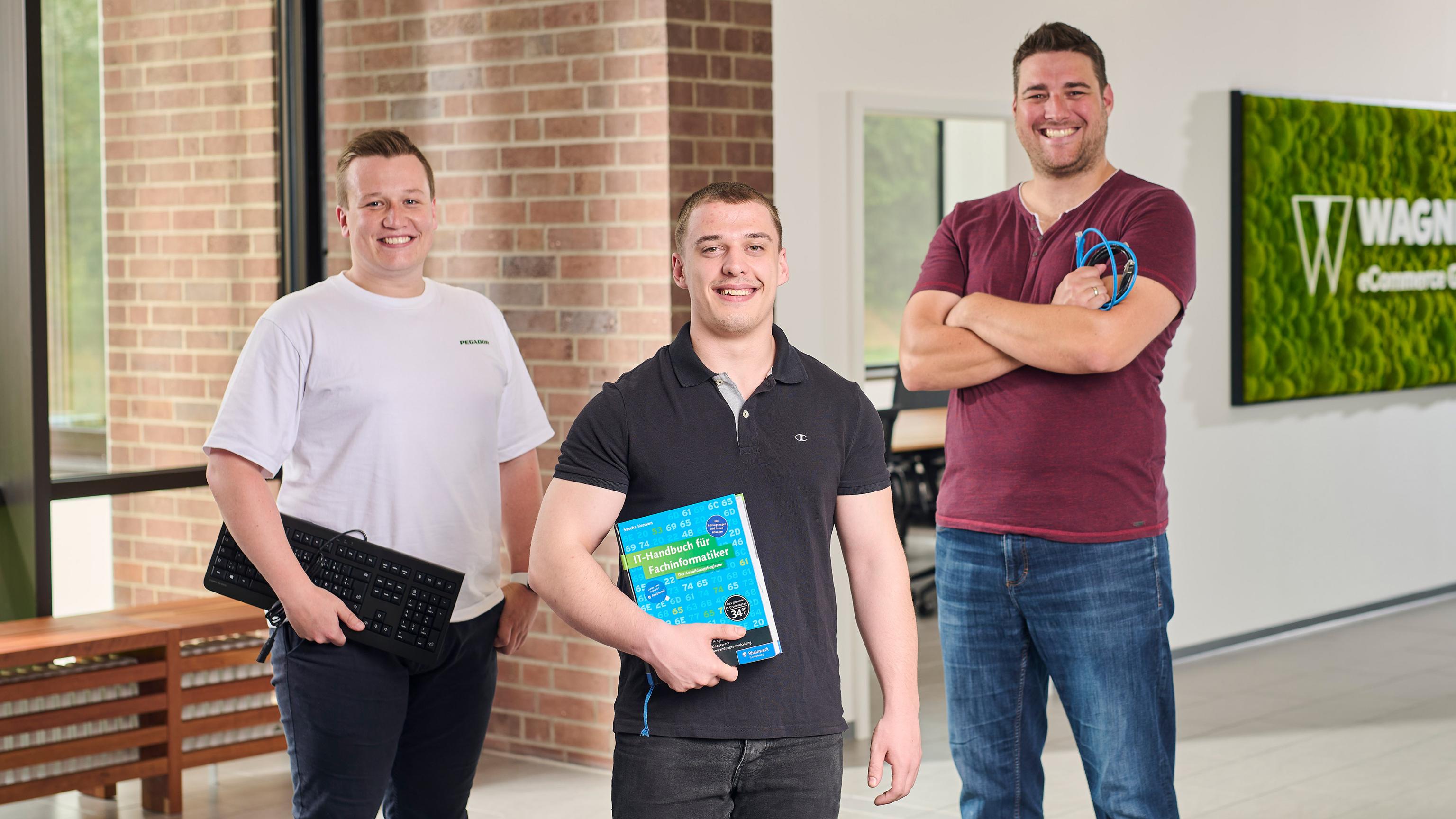 Das Bild zeigt drei Männer, die in einem modernen Bürogebäude vor einer Wand mit einem grünen Schild stehen, auf dem 'Wagner eCommerce Group' zu lesen ist. Der Mann links trägt ein weißes T-Shirt und hält eine Tastatur in der Hand. Der Mann in der Mitte trägt ein schwarzes Poloshirt und hält ein Buch mit dem Titel 'IT-Handbuch für Fachinformatiker'. Der Mann rechts trägt ein dunkelrotes T-Shirt und hält mehrere Kabel in der Hand. Alle drei Männer lächeln in die Kamera.
