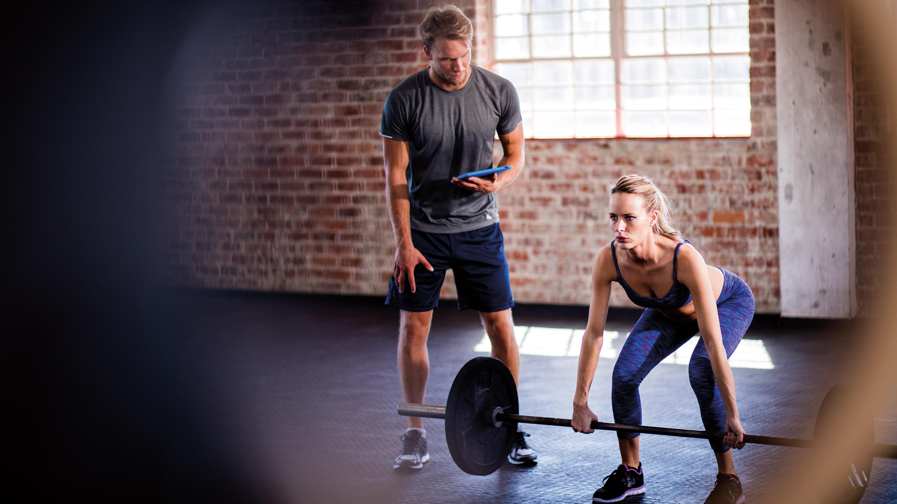 Trainer beobachtet eine Frau beim Gewichtheben in einem Fitnessstudio mit Backsteinwänden, beide konzentriert bei der Arbeit.