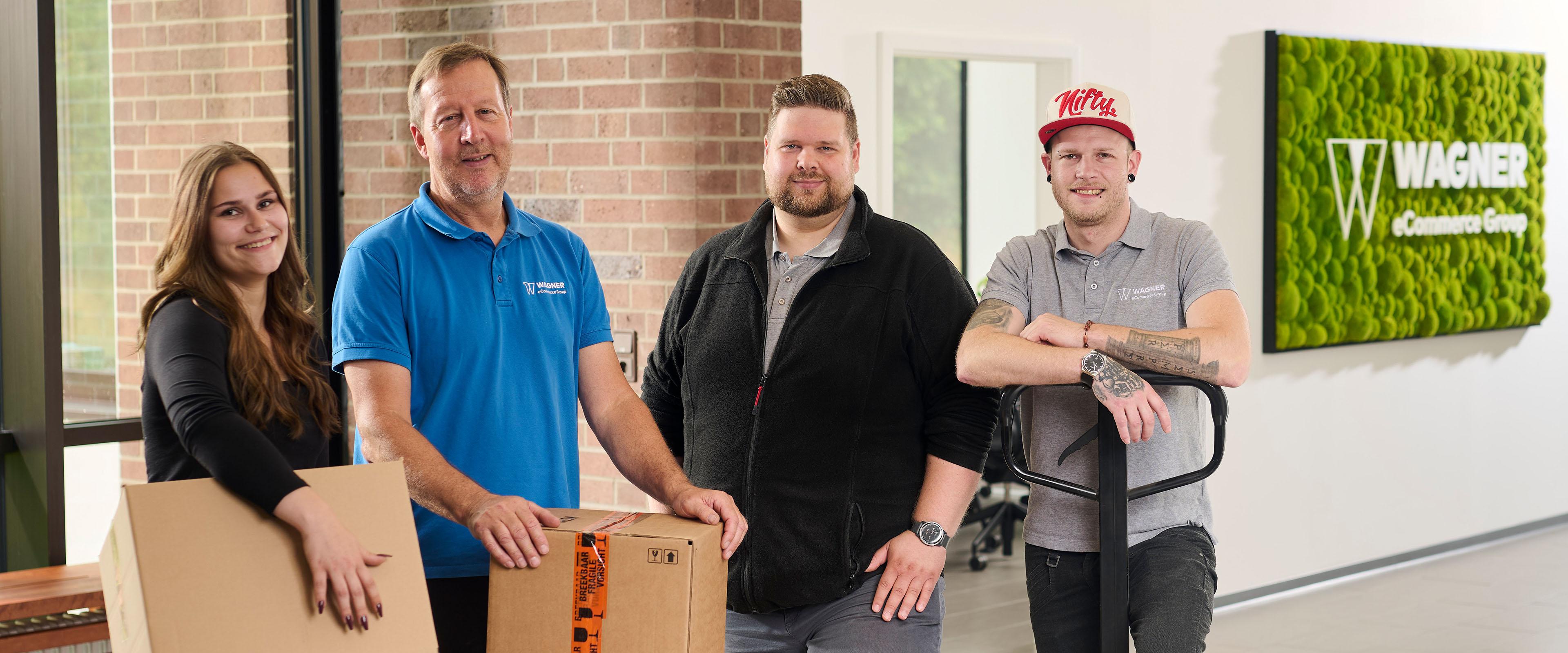 Das Bild zeigt vier Personen, die in einem modernen Bürogebäude vor einer Wand mit einem grünen Schild stehen, auf dem 'Wagner eCommerce Group' zu lesen ist. Links steht eine junge Frau mit langen braunen Haaren, die ein schwarzes Oberteil trägt und einen Karton in den Händen hält. Neben ihr steht ein älterer Mann in einem blauen Polo-Shirt mit dem Wagner-Logo, der ebenfalls einen Karton auf einem Tisch vor sich hält. Rechts von ihm steht ein Mann mit kurzem Haar und Bart, der eine schwarze Jacke trägt. Ganz rechts steht ein Mann in einem grauen Polo-Shirt mit dem Wagner-Logo und einer roten Baseballkappe, der einen Palettenhubwagen hält. Alle Personen lächeln in die Kamera.