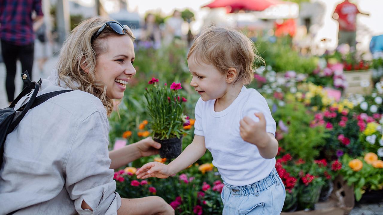 Lächelnde Frau zeigt einem kleinen Kind Blumen auf einem bunten Markt, beide wirken glücklich und entspannt.