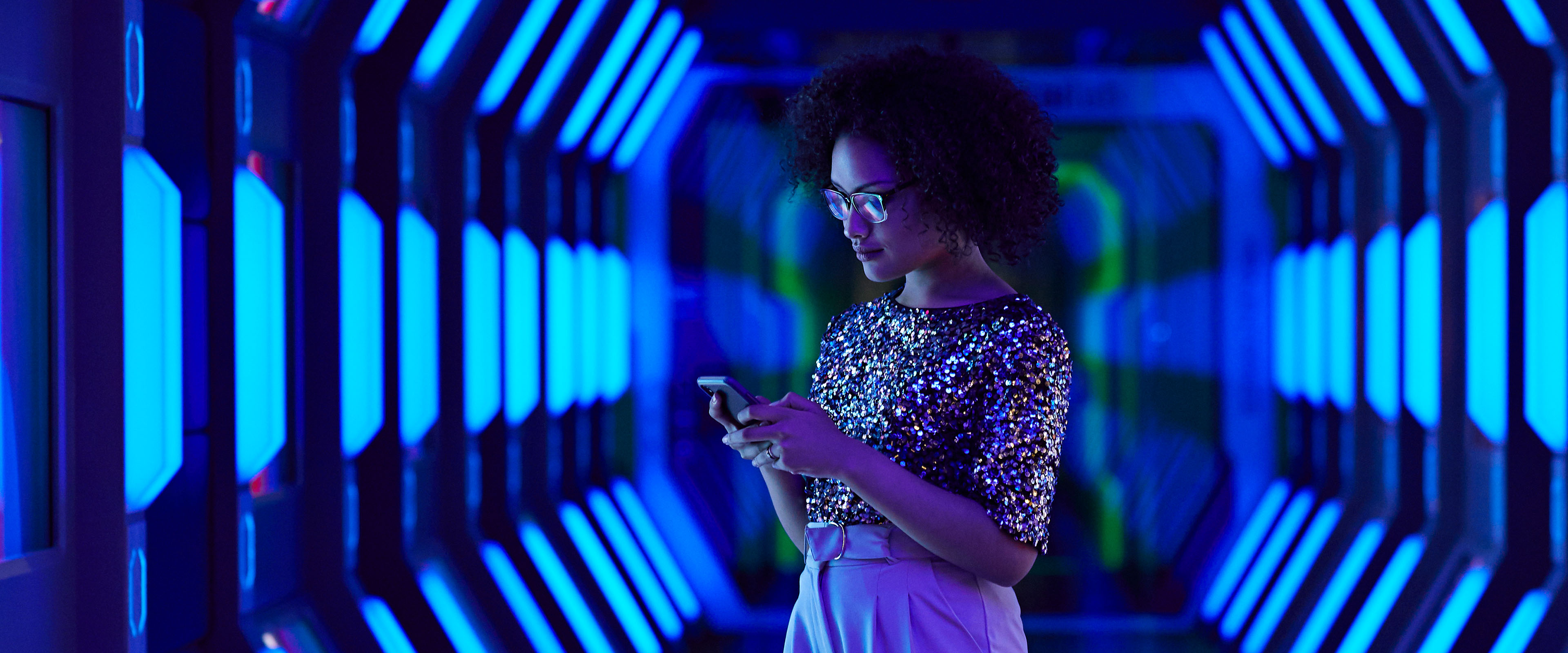 Frau in glitzerndem Oberteil steht in einem futuristisch beleuchteten Korridor und schaut auf ihr Smartphone.