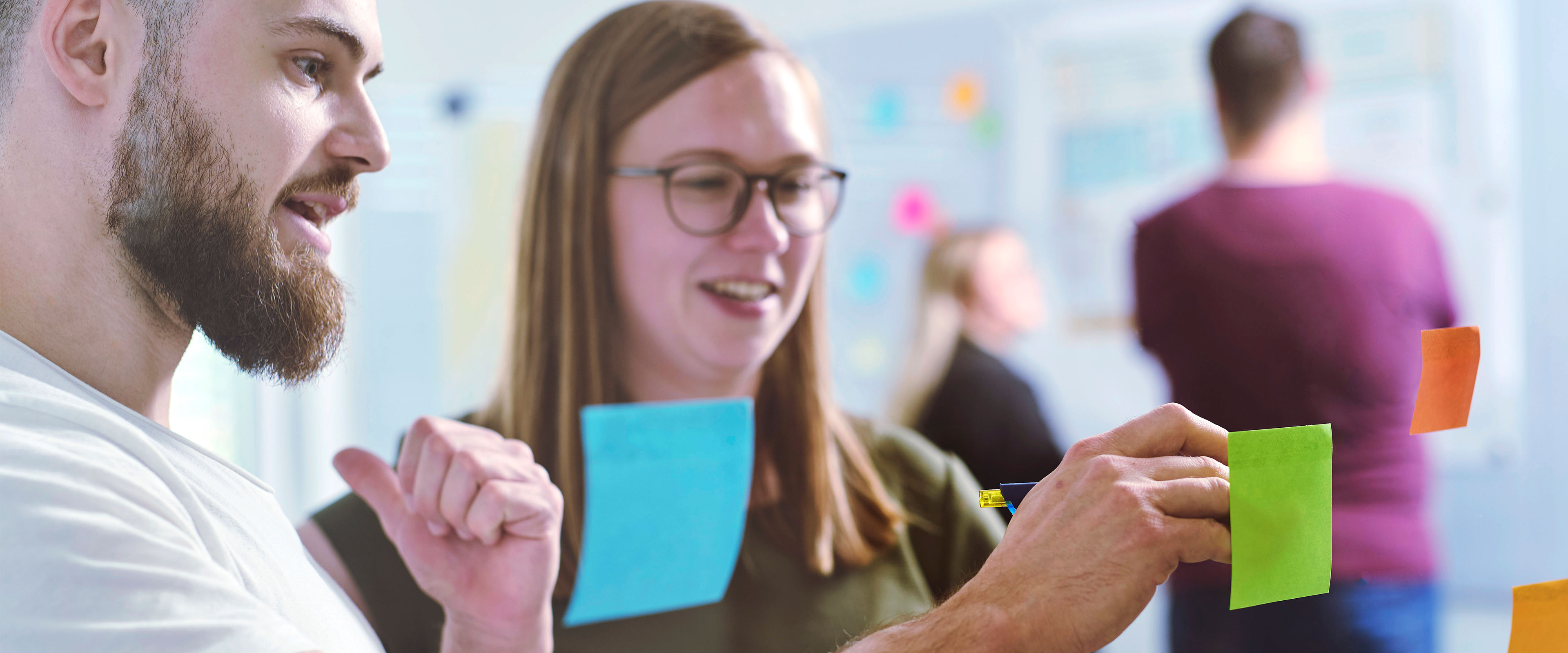 Zwei Personen diskutieren und kleben bunte Haftnotizen an eine Glaswand, während im Hintergrund andere arbeiten.