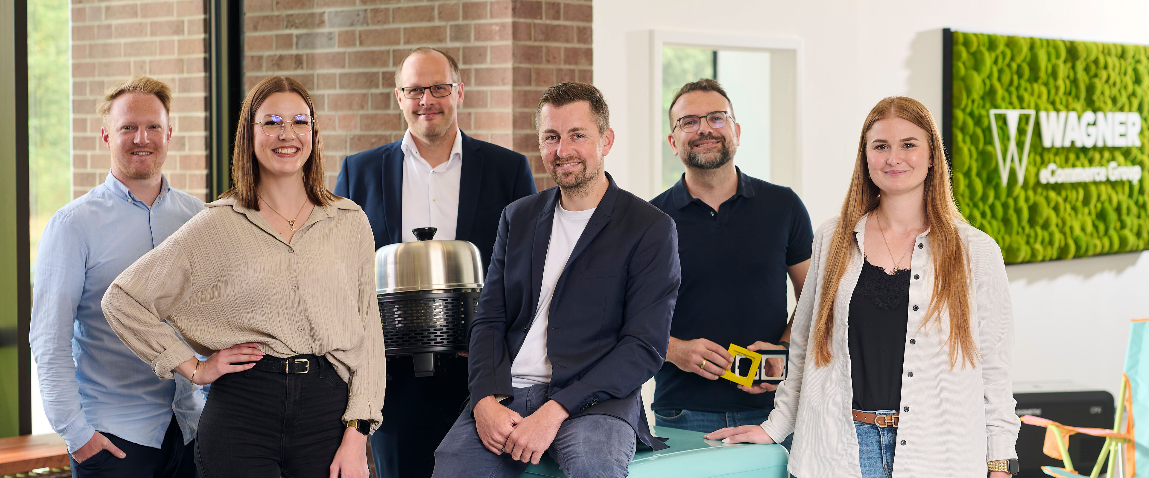 Fünf Personen stehen in einem Büro und halten jeweils eine Nummernkarte von 1 bis 4 in der Hand. Im Hintergrund ist ein Whiteboard mit Zahlen zu sehen. Das Büro ist modern gestaltet und hat eine grüne Pflanzenwand.