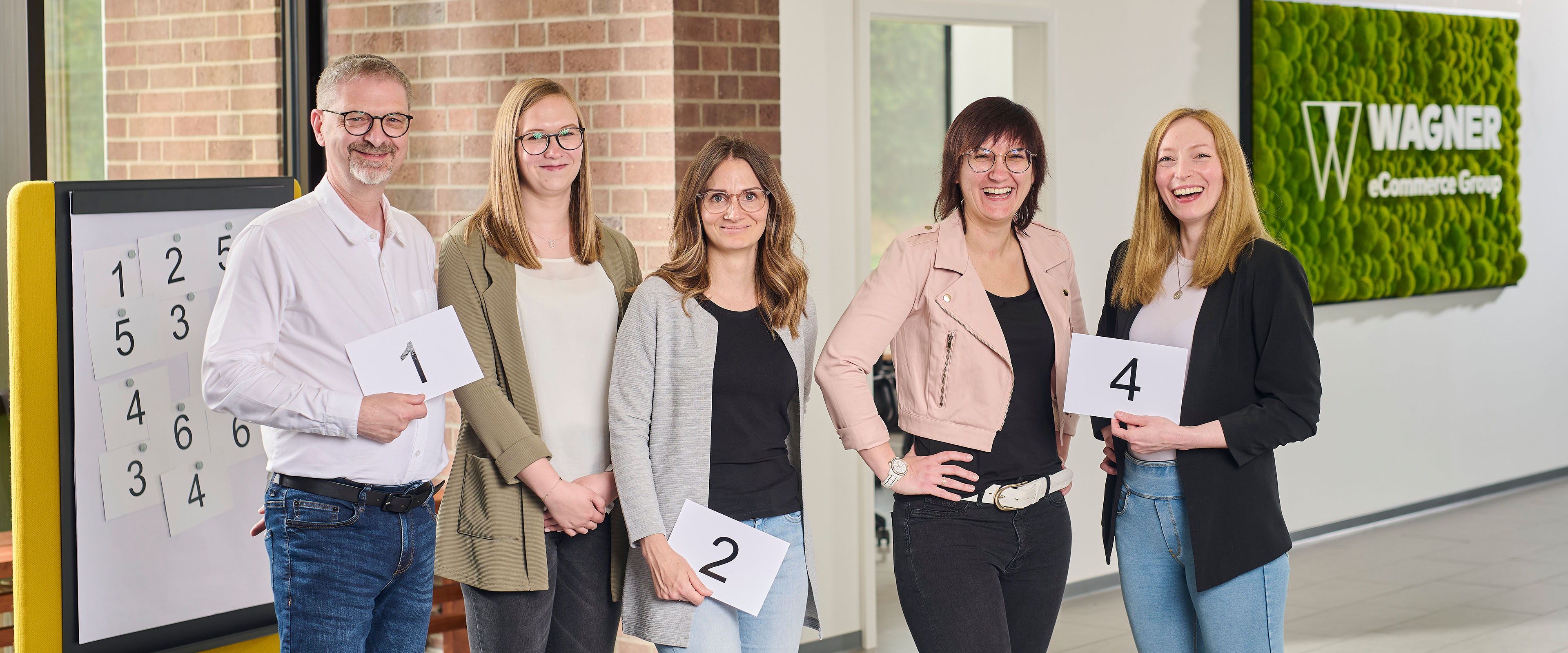 Ein Team von fünf Personen posiert mit nummerierten Karten in einem hellen Büro. Ein Zahlenbrett und ein grünes Wandschild mit der Aufschrift 'Wagner eCommerce Group' sind im Hintergrund sichtbar.