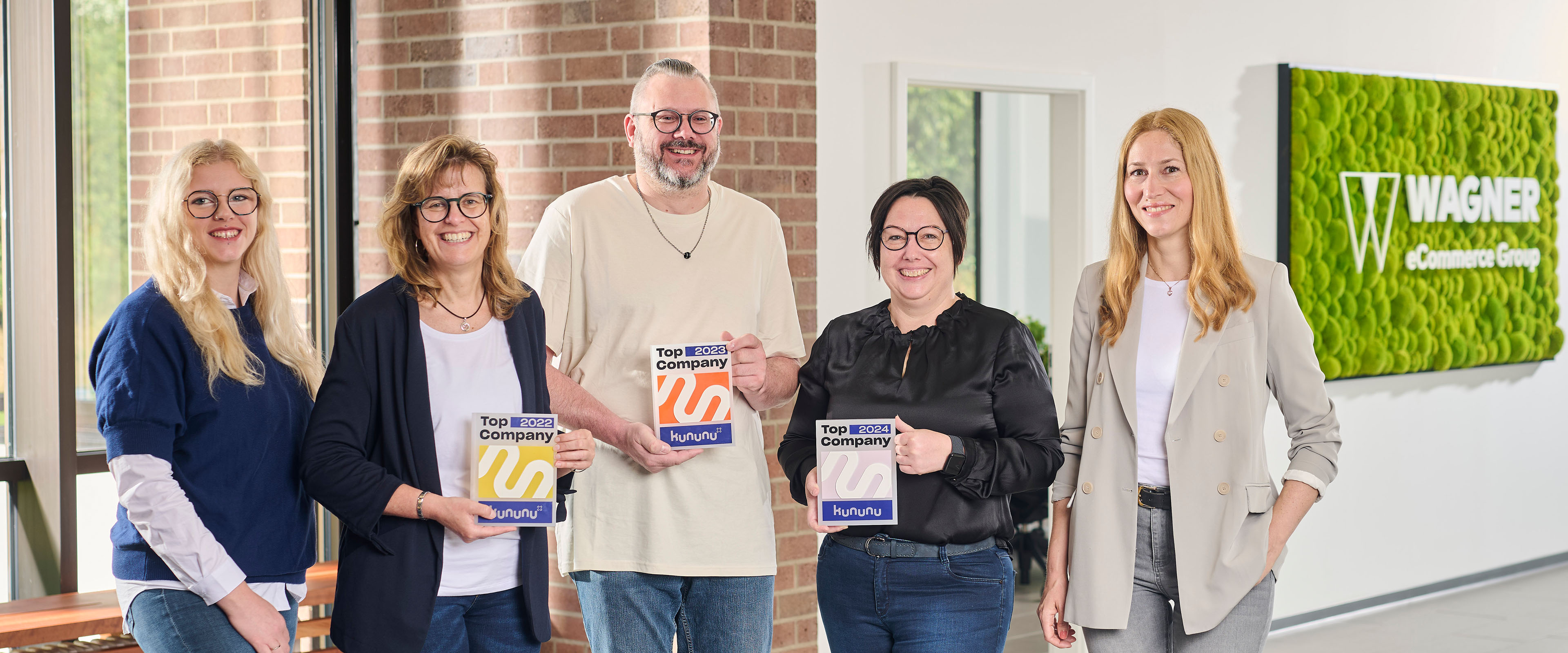 Fünf Menschen stehen in einem modernen Bürogebäude. Zwei von ihnen sind mit der Auszeichnung „Top Company“ ausgezeichnet. An der Wand hängt ein grünes Schild mit der Aufschrift „Wagner Group“. Sie lächeln und posieren neben großen Fenstern und einem grünen Logoschild.