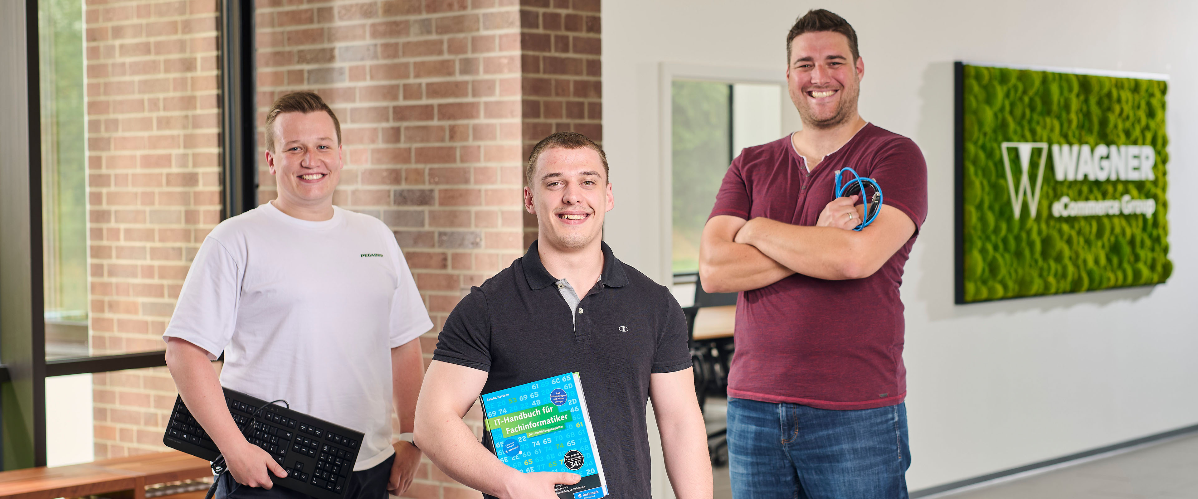 Das Bild zeigt drei Männer, die in einem modernen Bürogebäude vor einer Wand mit einem grünen Schild stehen, auf dem 'Wagner eCommerce Group' zu lesen ist. Der Mann links trägt ein weißes T-Shirt und hält eine Tastatur in der Hand. Der Mann in der Mitte trägt ein schwarzes Poloshirt und hält ein Buch mit dem Titel 'IT-Handbuch für Fachinformatiker'. Der Mann rechts trägt ein dunkelrotes T-Shirt und hält mehrere Kabel in der Hand. Alle drei Männer lächeln in die Kamera.