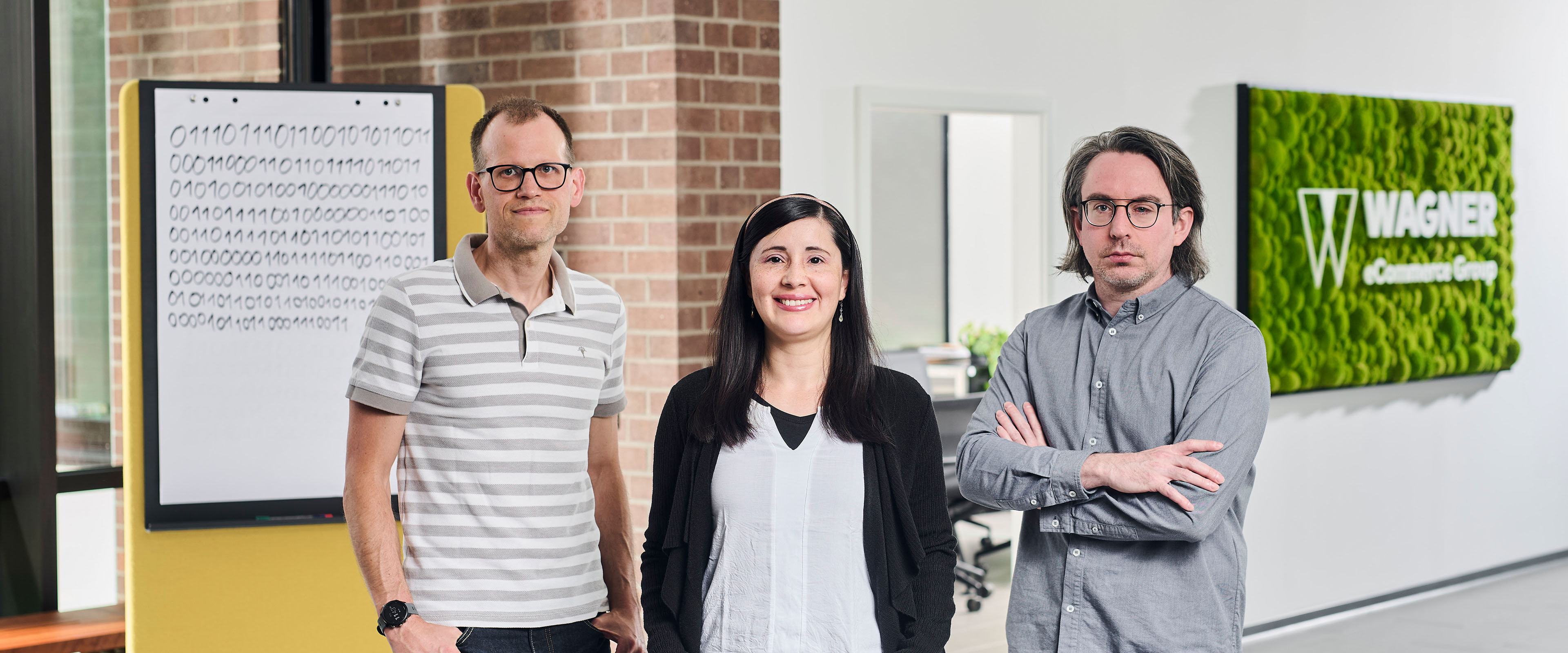 Das Bild zeigt drei Personen, die in einem modernen Bürogebäude vor einer Wand mit einem grünen Schild stehen, auf dem 'Wagner eCommerce Group' zu lesen ist. Links steht ein Mann mit Brille und einem grau-weiß gestreiften Poloshirt, der seine Hände in den Hosentaschen hat. In der Mitte steht eine Frau mit langen dunklen Haaren, einem weißen Oberteil und einer schwarzen Strickjacke, die in die Kamera lächelt. Rechts steht ein Mann mit Brille und schulterlangem Haar, der ein graues Hemd trägt und die Arme verschränkt hat. Im Hintergrund ist ein Whiteboard mit Binärcode-Muster zu sehen.