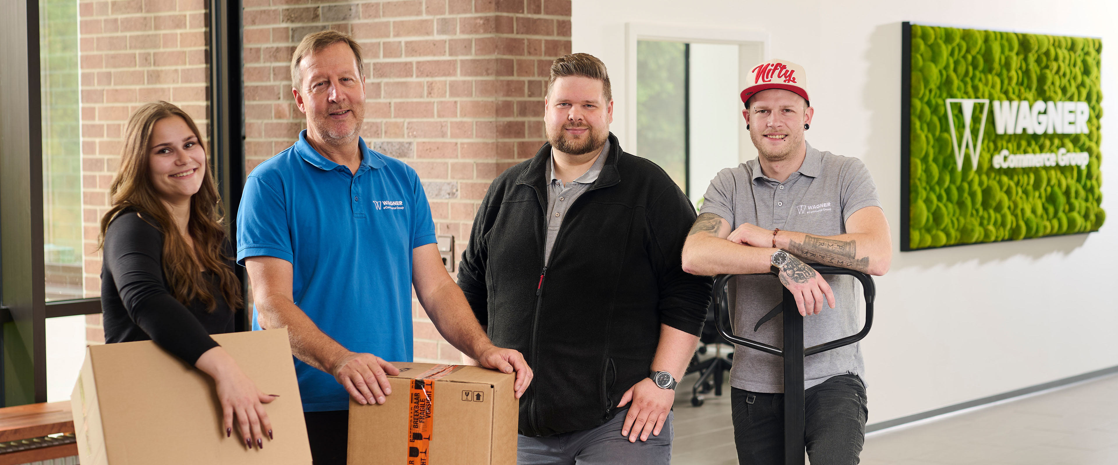 Das Bild zeigt vier Personen, die in einem modernen Bürogebäude vor einer Wand mit einem grünen Schild stehen, auf dem 'Wagner eCommerce Group' zu lesen ist. Links steht eine junge Frau mit langen braunen Haaren, die ein schwarzes Oberteil trägt und einen Karton in den Händen hält. Neben ihr steht ein älterer Mann in einem blauen Polo-Shirt mit dem Wagner-Logo, der ebenfalls einen Karton auf einem Tisch vor sich hält. Rechts von ihm steht ein Mann mit kurzem Haar und Bart, der eine schwarze Jacke trägt. Ganz rechts steht ein Mann in einem grauen Polo-Shirt mit dem Wagner-Logo und einer roten Baseballkappe, der einen Palettenhubwagen hält. Alle Personen lächeln in die Kamera.