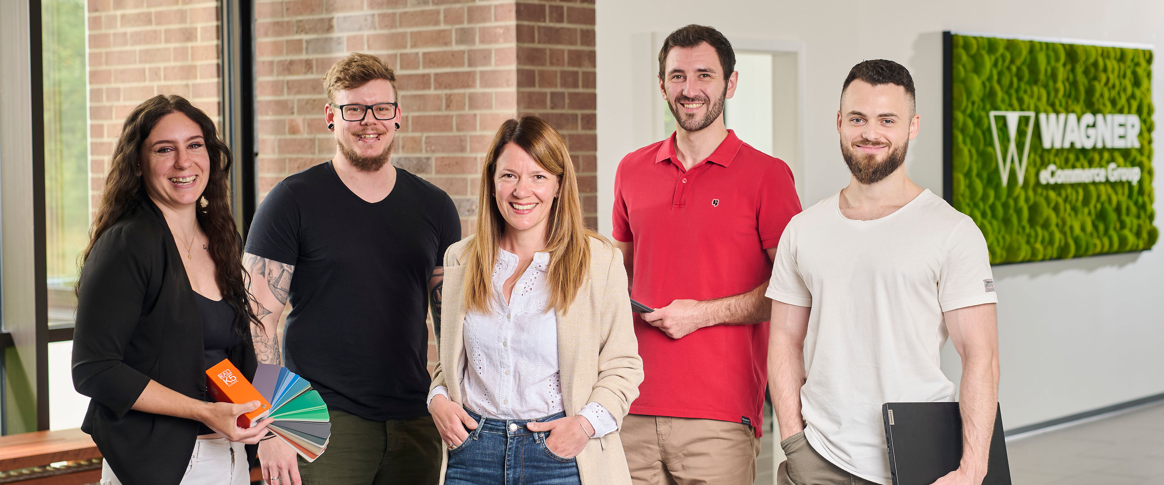 Das Bild zeigt fünf Personen, die in einem modernen Bürogebäude vor einer Wand mit einem grünen Schild stehen, auf dem 'Wagner eCommerce Group' zu lesen ist. Links steht eine Frau mit lockigem braunem Haar, die ein schwarzes Oberteil und eine weiße Hose trägt. Sie hält Farbpaletten in der Hand. Neben ihr steht ein Mann mit Brille und kurzem Haar, der ein schwarzes T-Shirt und olivgrüne Shorts trägt. In der Mitte steht eine Frau mit langen braunen Haaren, die ein beiges Sakko, ein weißes Hemd und eine blaue Jeans trägt. Rechts von ihr steht ein Mann mit einem roten Poloshirt, der ein Tablet in der Hand hält. Ganz rechts steht ein Mann mit kurzem Haar und Bart, der ein weißes T-Shirt und khakifarbene Hosen trägt und einen Laptop in der Hand hält. Alle Personen lächeln in die Kamera