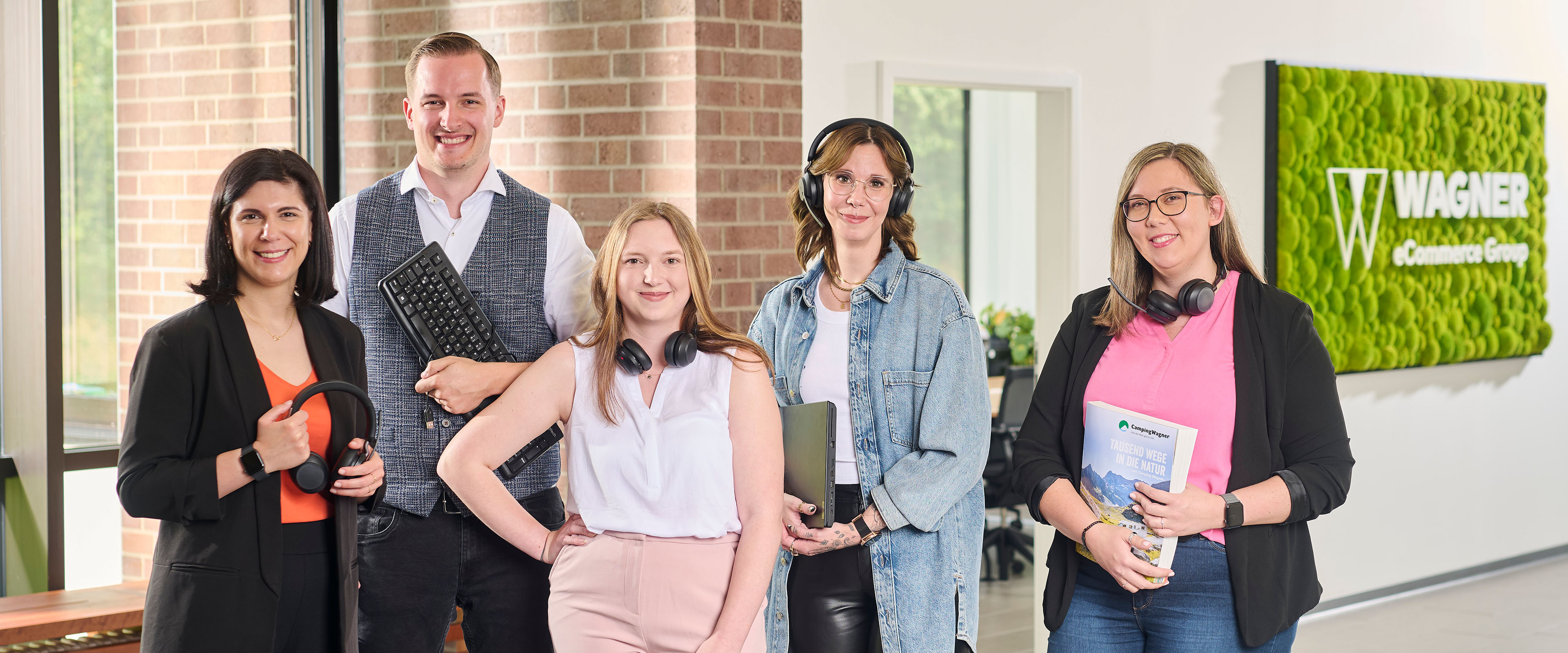 Fünf lächelnde Personen in einem Büro, halten Kopfhörer, Tastatur, Laptop und Magazin, vor einem Wagner eCommerce Schild.