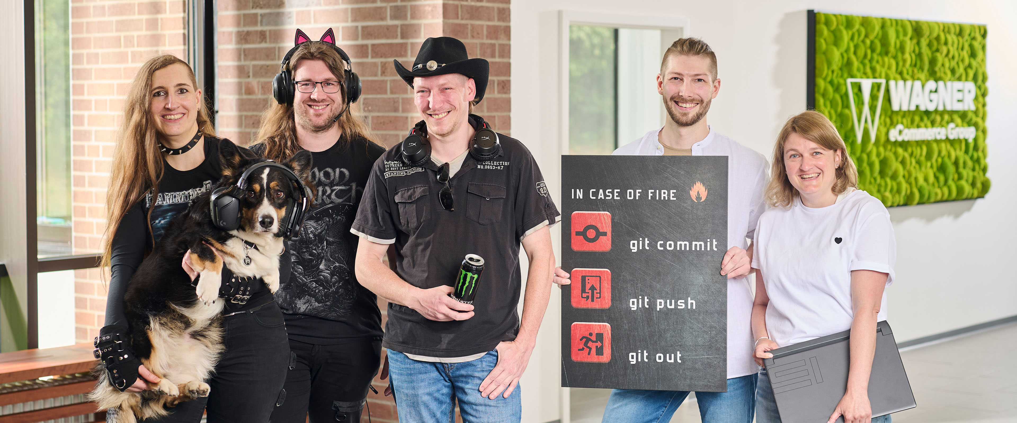 Das Bild zeigt sechs Personen, die in einem modernen Bürogebäude vor einer Wand mit einem grünen Schild stehen, auf dem 'Wagner eCommerce Group' zu lesen ist. Links steht eine Frau mit langen braunen Haaren, die ein schwarzes Metal-T-Shirt trägt und einen Hund hält, der Kopfhörer trägt. Neben ihr steht ein Mann mit langen Haaren und einer Katzenohren-Kopfbedeckung, der ebenfalls ein schwarzes T-Shirt trägt. Daneben steht ein Mann mit Cowboyhut und Kopfhörern um den Hals, der eine Dose Monster-Energy in der Hand hält. Rechts von ihm steht ein Mann, der ein Schild mit der Aufschrift 'In Case of Fire: git commit, git push, git out' hält. Ganz rechts steht eine Frau mit blonden Haaren und weißem T-Shirt, die ein Laptop unter dem Arm hält. Alle Personen lächeln in die Kamera.
