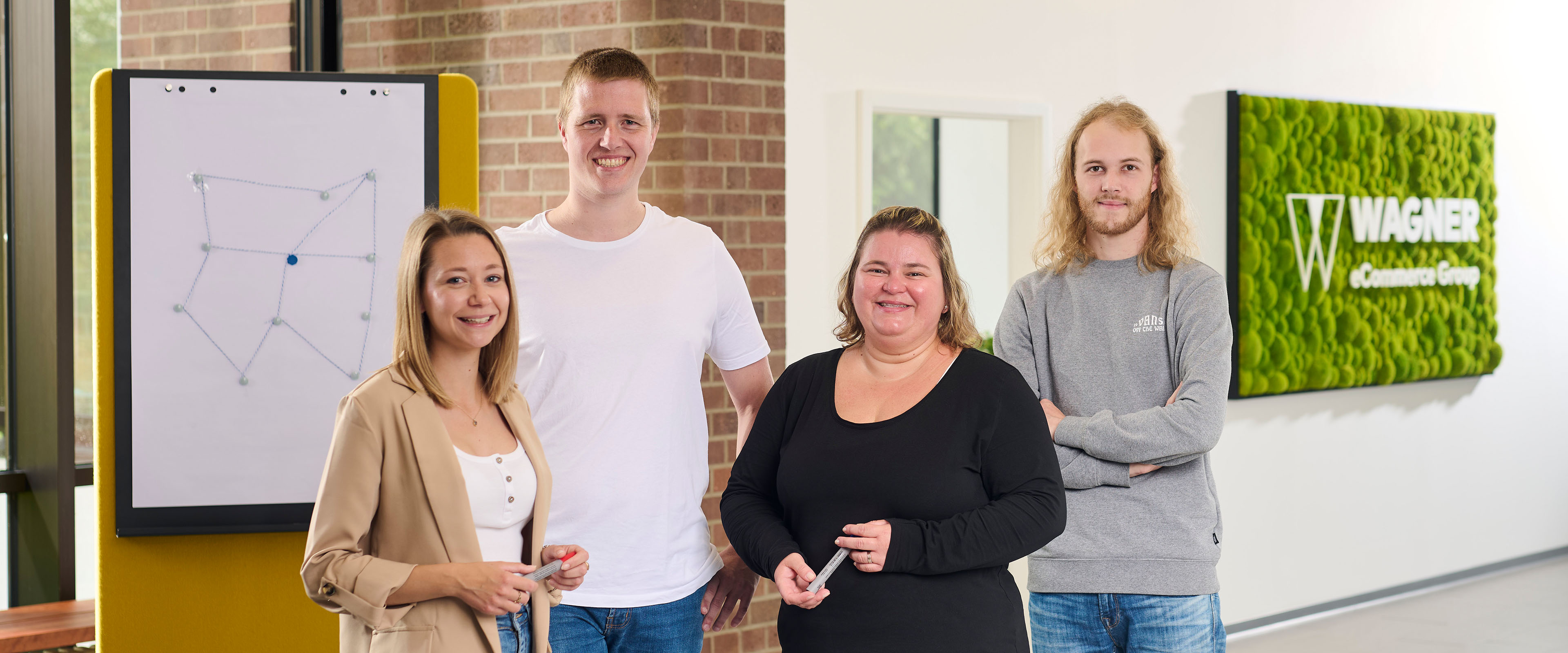 Eine Gruppe von vier lächelnden Personen steht in einem modernen Büro. Sie befinden sich vor einem Whiteboard mit einer einfachen geometrischen Zeichnung und einem grünen Wandschild, auf dem 'Wagner eCommerce Group' steht.