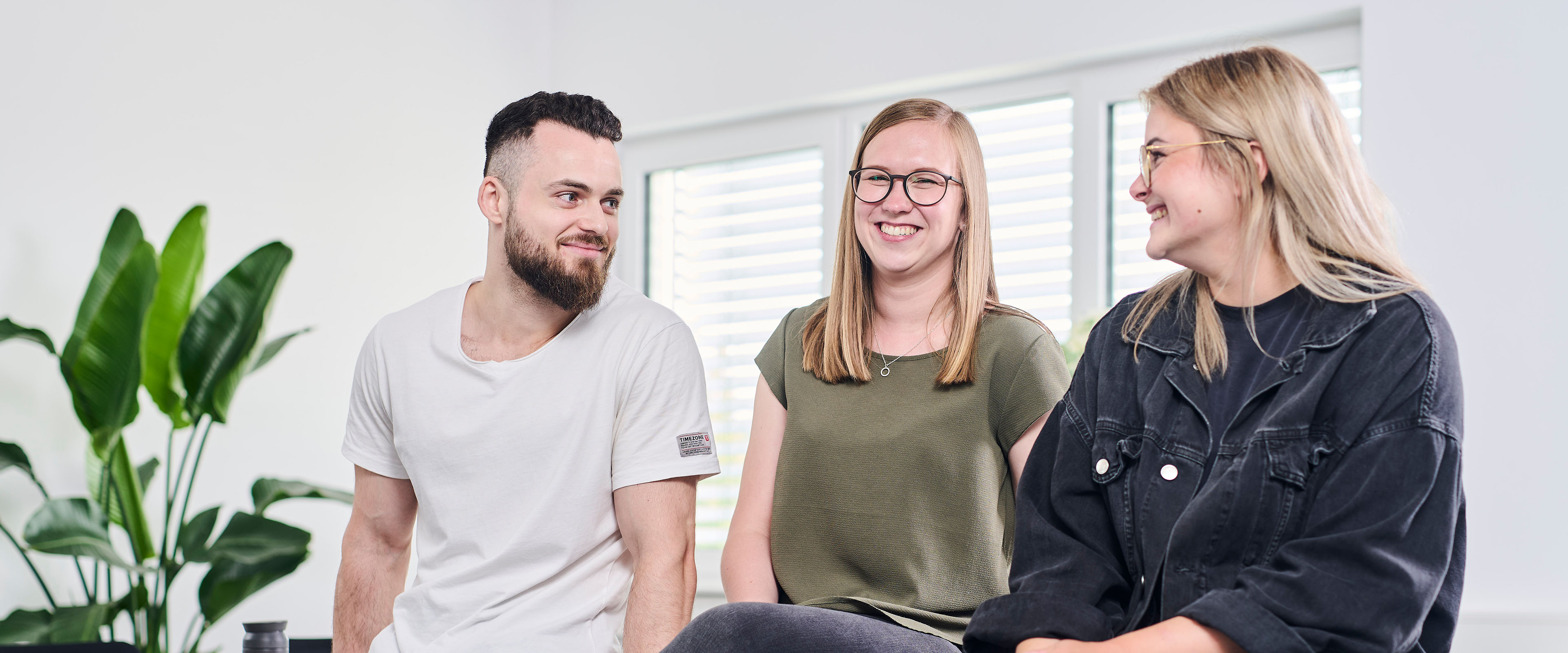 Drei lächelnde Personen sitzen auf einem Tisch in einem hellen Raum, sie unterhalten sich entspannt miteinander.