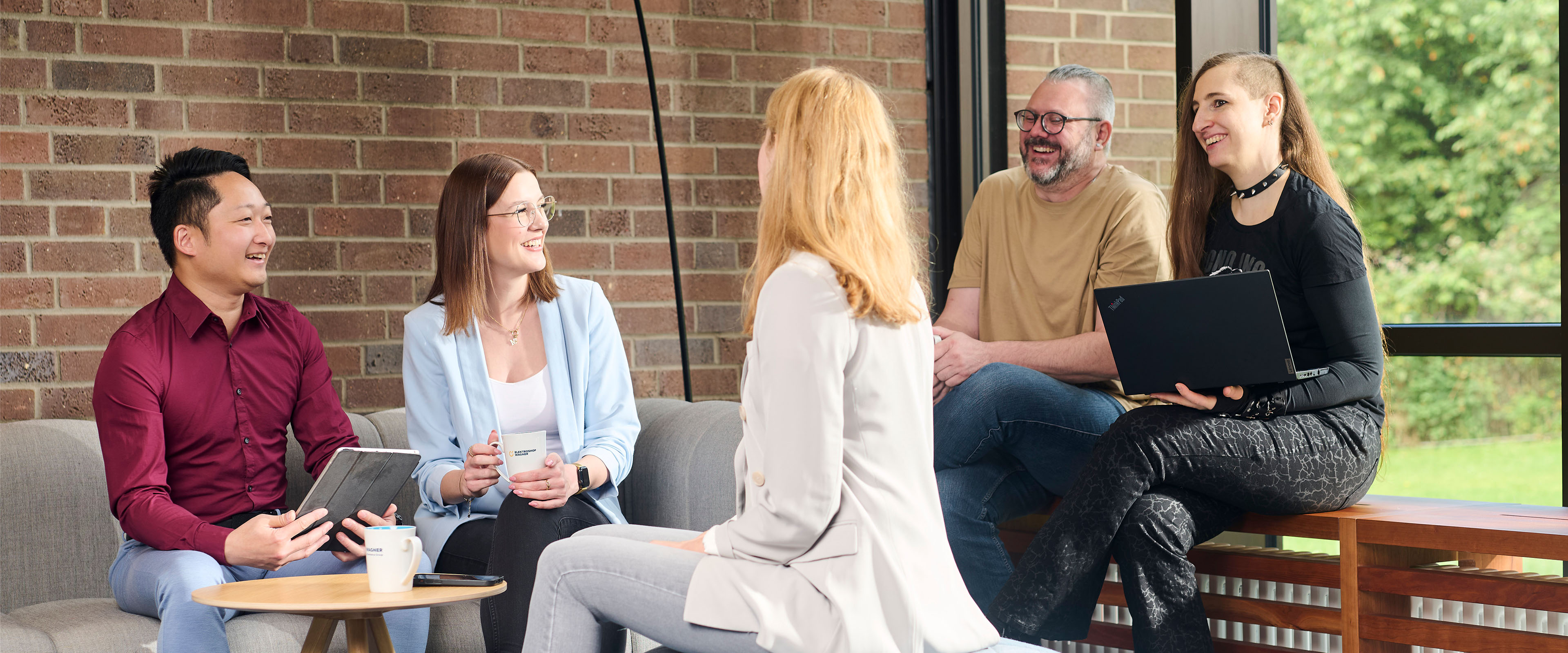 Fünf Personen sitzen in einer entspannten Runde in einem modernen Büro, lächelnd und im Gespräch vertieft.