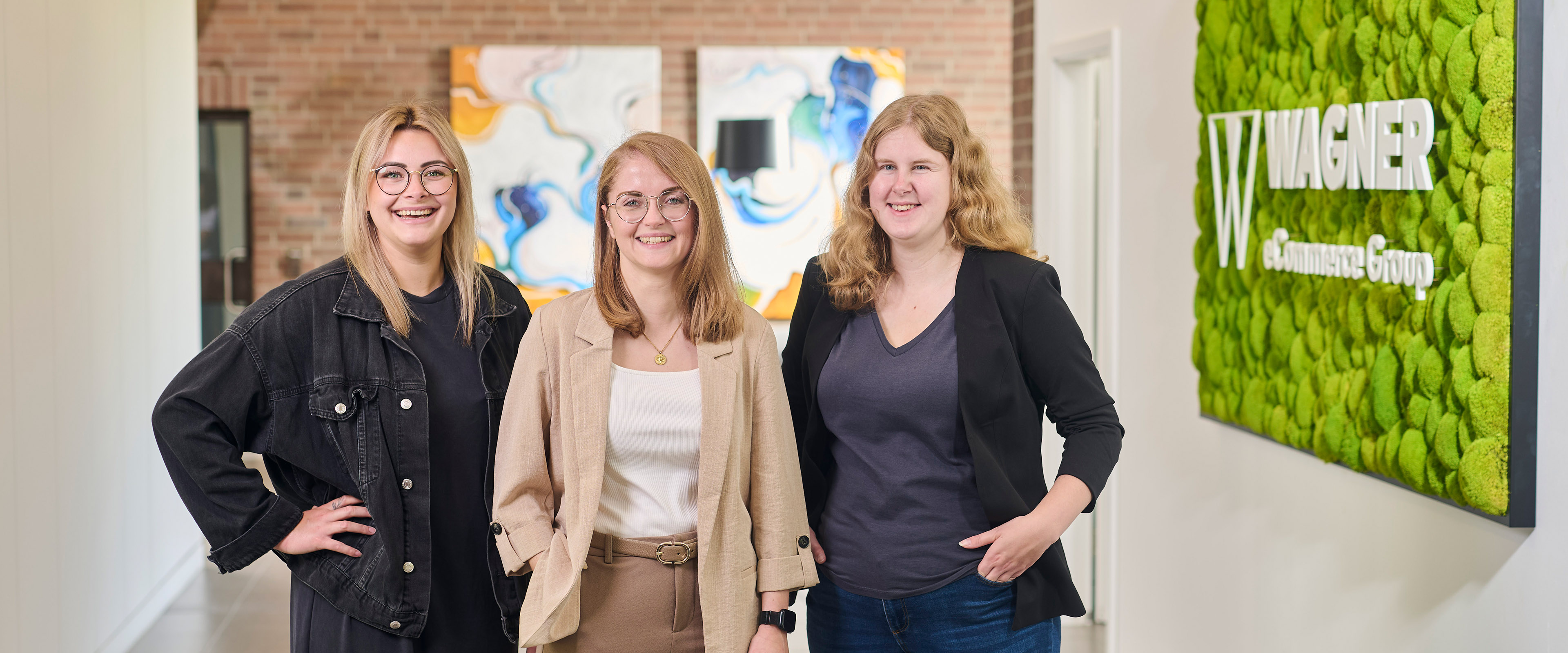 Drei lächelnde Frauen stehen in einem Flur vor einem Wagner eCommerce Group Schild, hinter ihnen bunte Wandkunst.