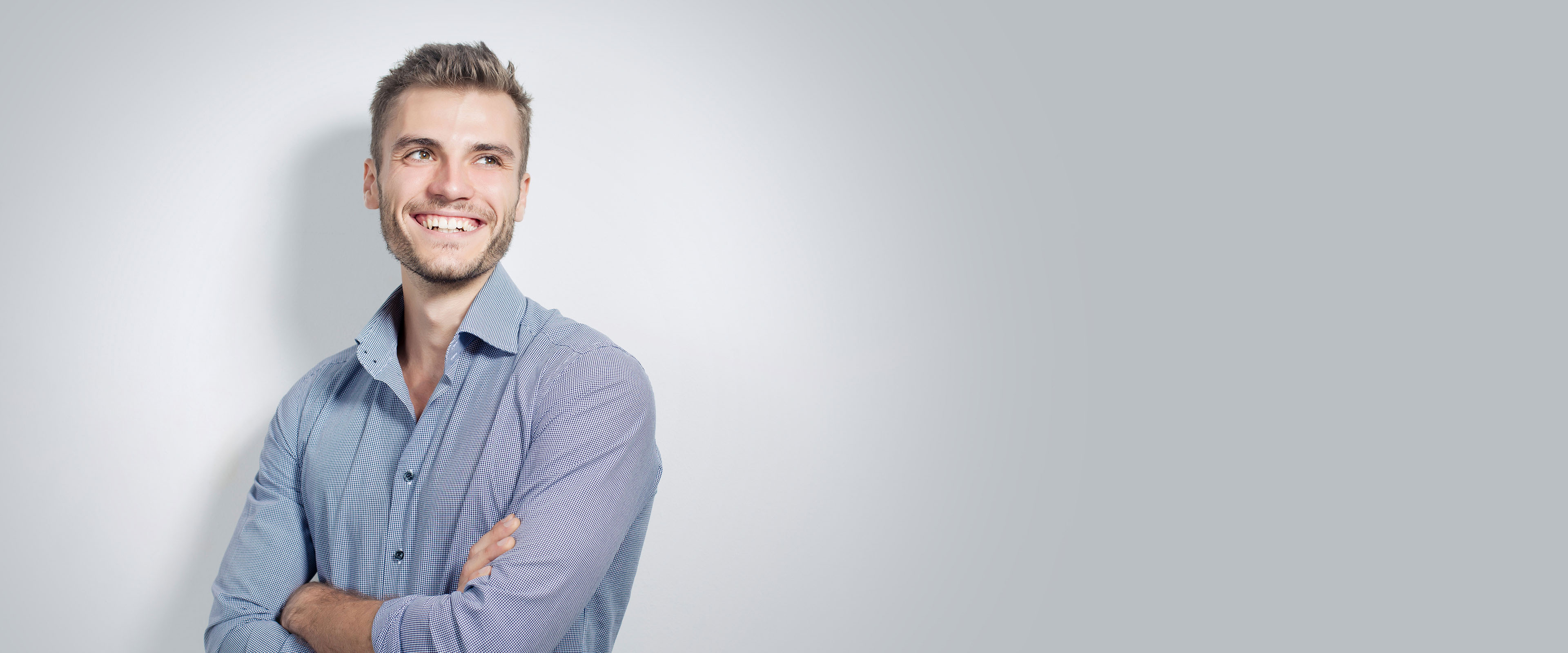 Lächelnder Mann in einem blauen Hemd steht mit verschränkten Armen vor einem hellen Hintergrund, blickt zur Seite.