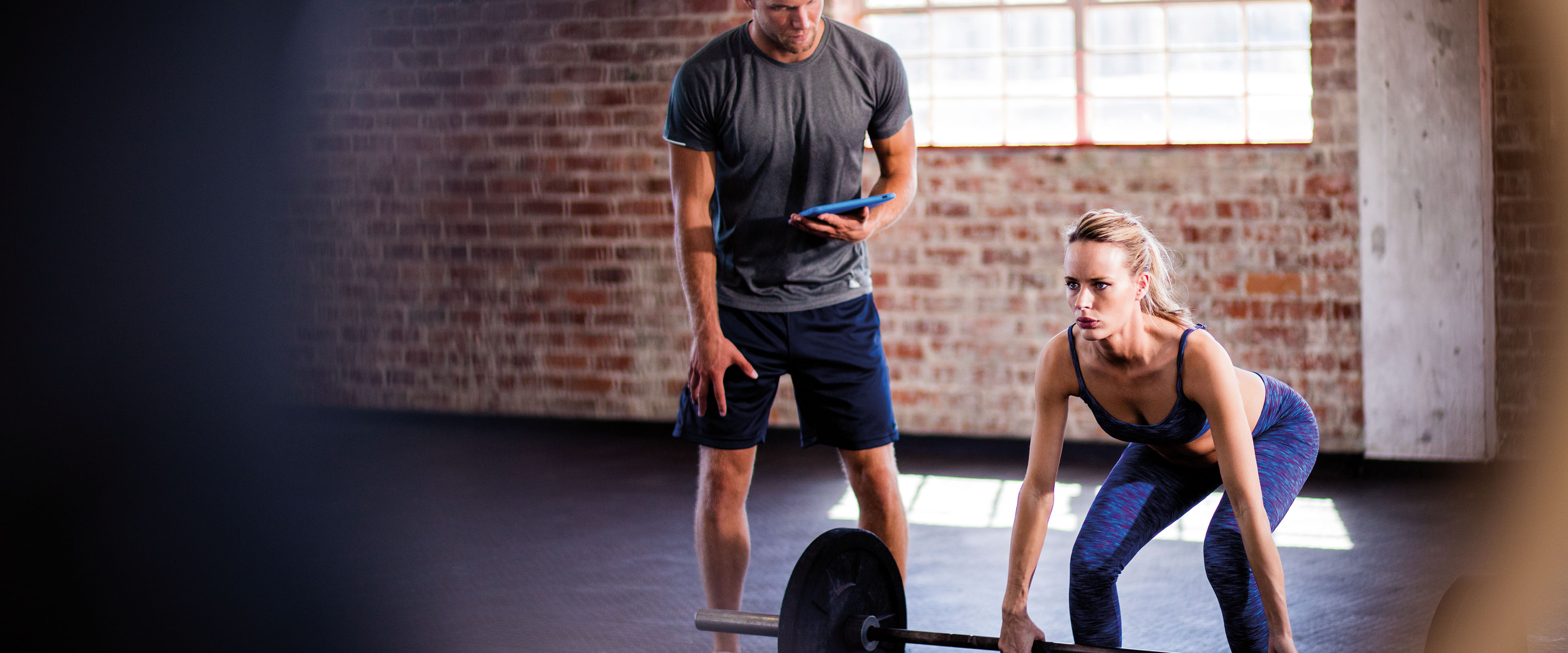 Trainer beobachtet eine Frau beim Gewichtheben in einem Fitnessstudio mit Backsteinwänden, beide konzentriert bei der Arbeit.