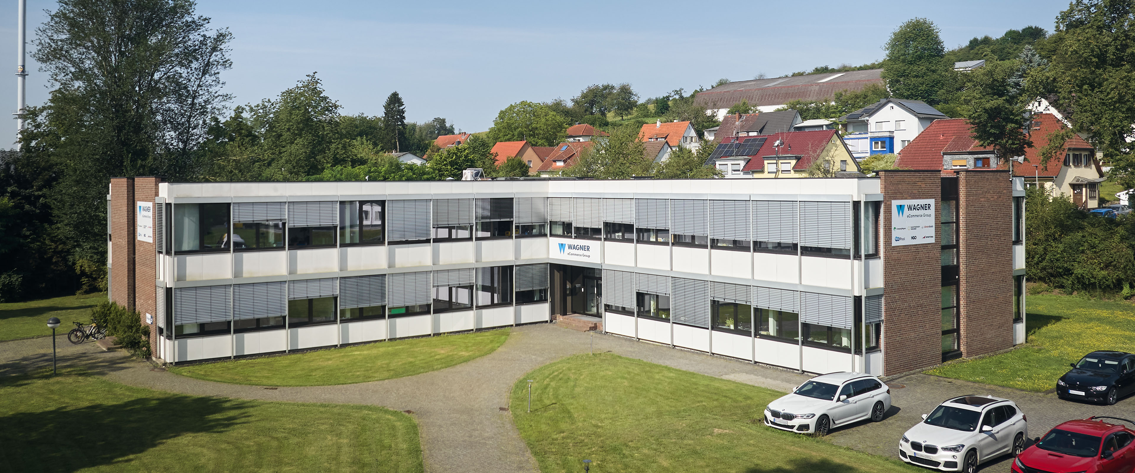 Außenansicht eines modernen Bürogebäudes der Wagner eCommerce Group, umgeben von Rasenflächen und parkenden Autos.