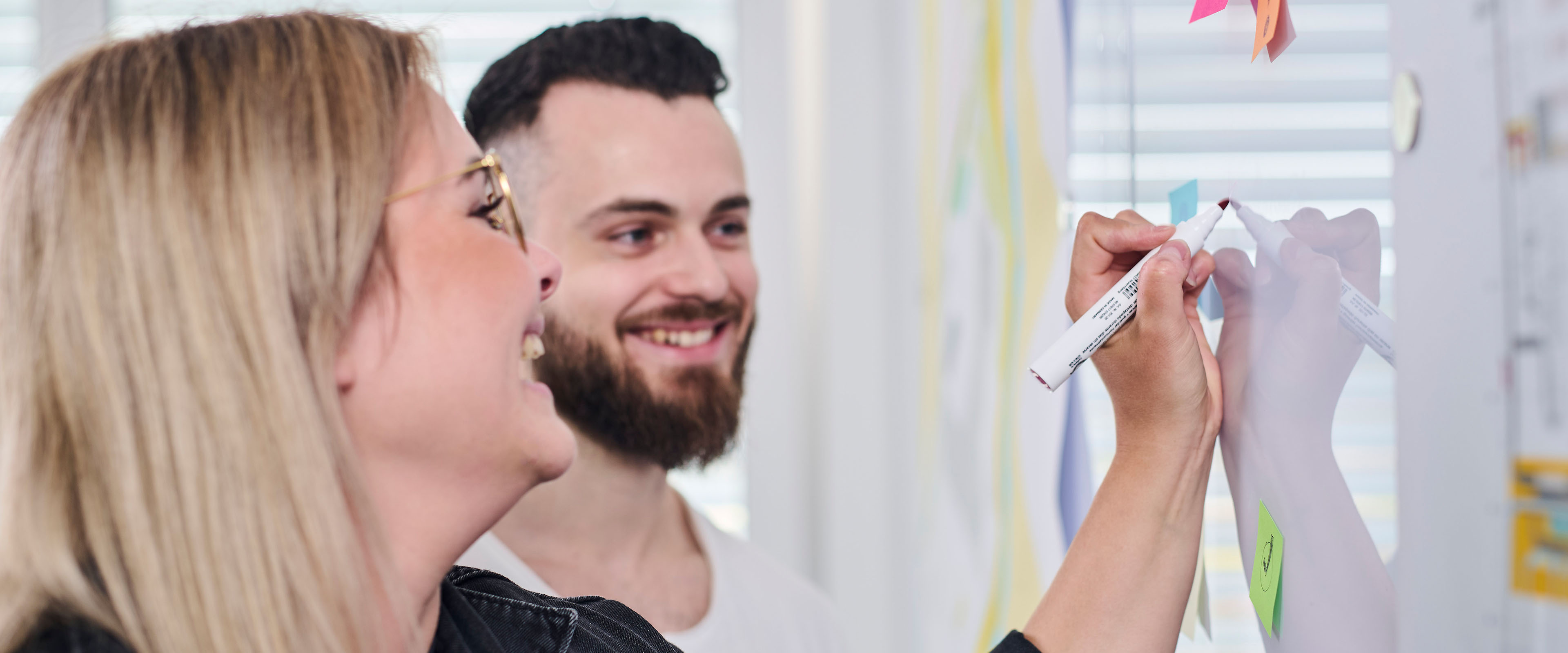 Zwei Personen lächeln, während sie gemeinsam an einer Glaswand mit Stift und Haftnotizen arbeiten.