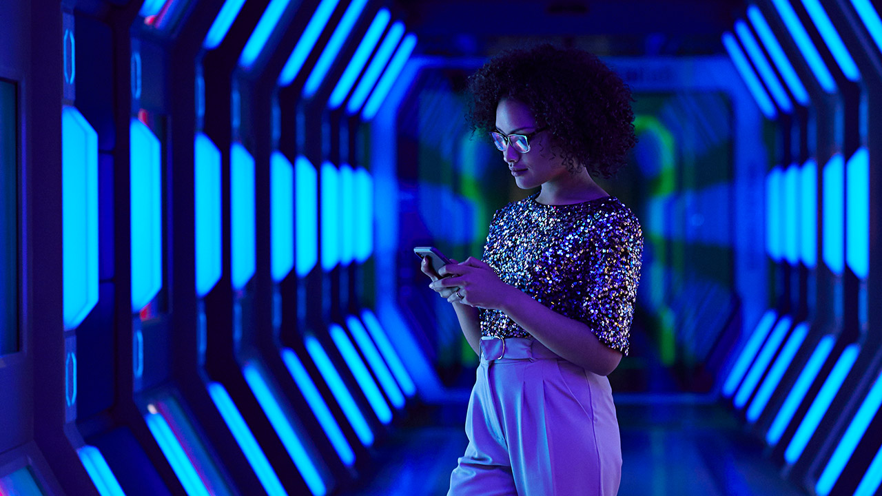 Frau in glitzerndem Oberteil steht in einem futuristisch beleuchteten Korridor und schaut auf ihr Smartphone.