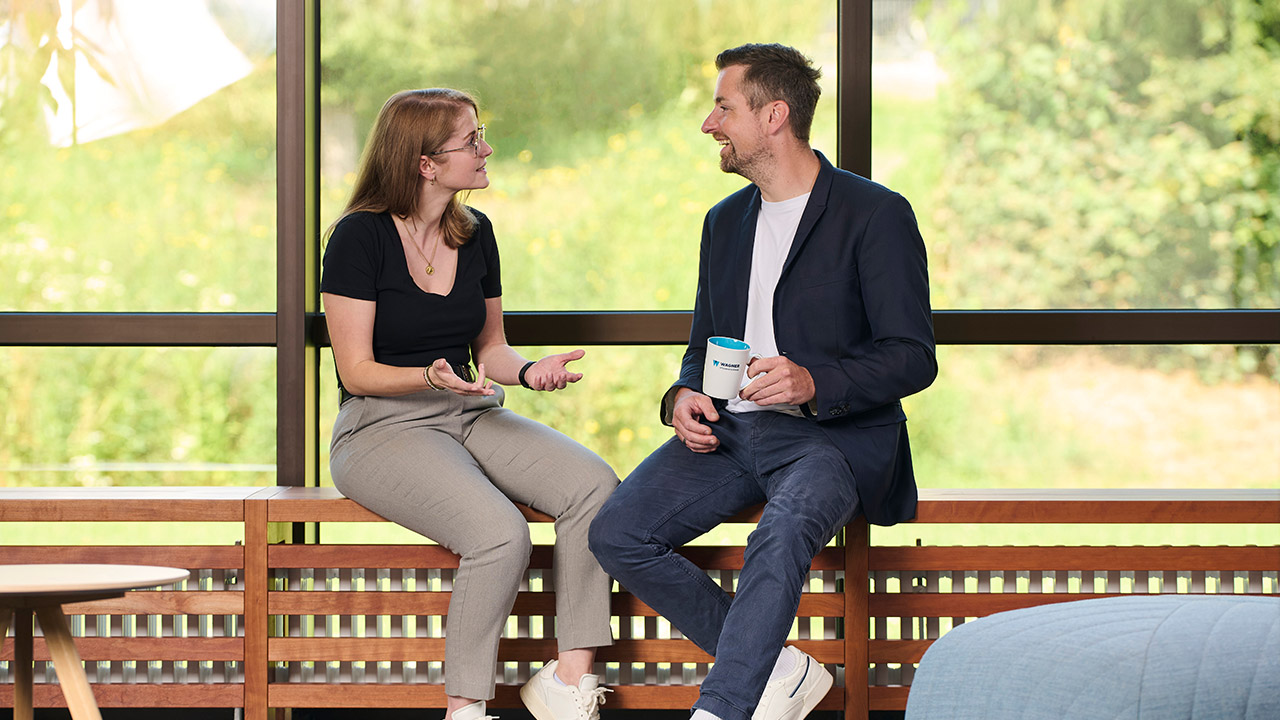 Zwei Personen sitzen und unterhalten sich, eine Frau in Schwarz und ein Mann im Anzug, mit einer Tasse in der Hand.