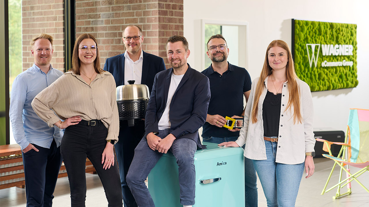 Fünf Personen stehen in einem Büro und halten jeweils eine Nummernkarte von 1 bis 4 in der Hand. Im Hintergrund ist ein Whiteboard mit Zahlen zu sehen. Das Büro ist modern gestaltet und hat eine grüne Pflanzenwand.