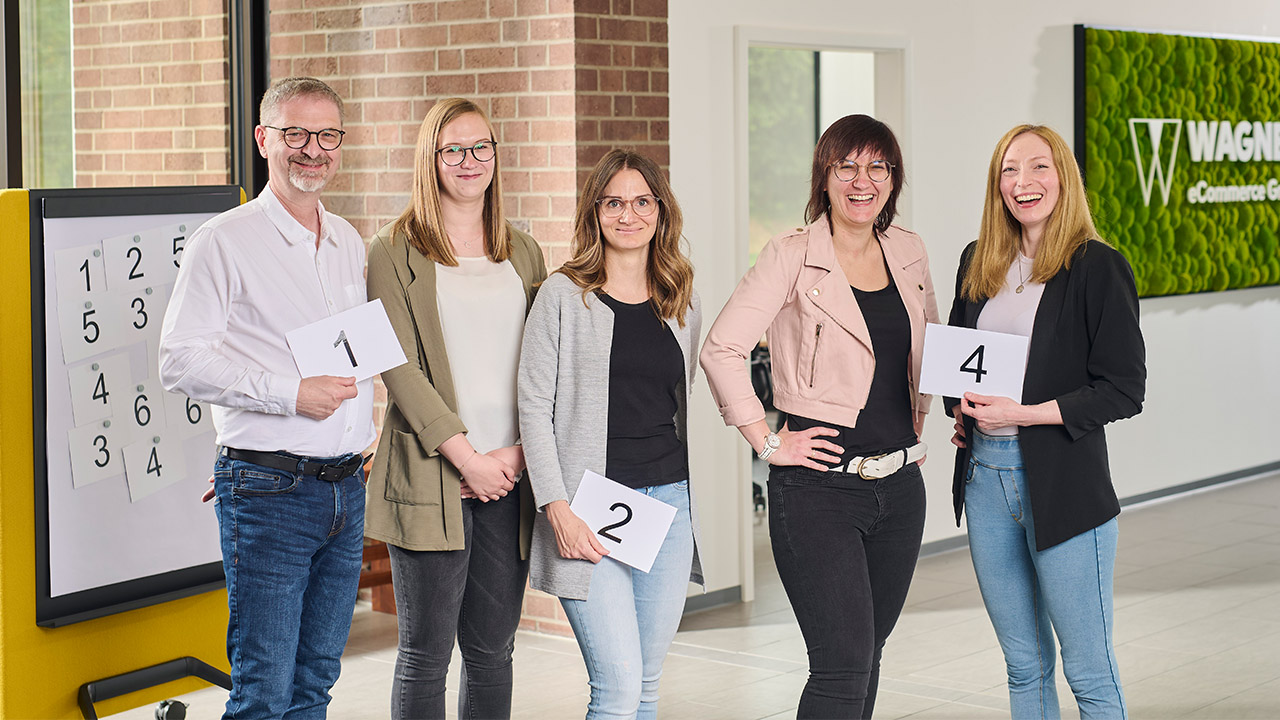 Ein Team von fünf Personen posiert mit nummerierten Karten in einem hellen Büro. Ein Zahlenbrett und ein grünes Wandschild mit der Aufschrift 'Wagner eCommerce Group' sind im Hintergrund sichtbar.