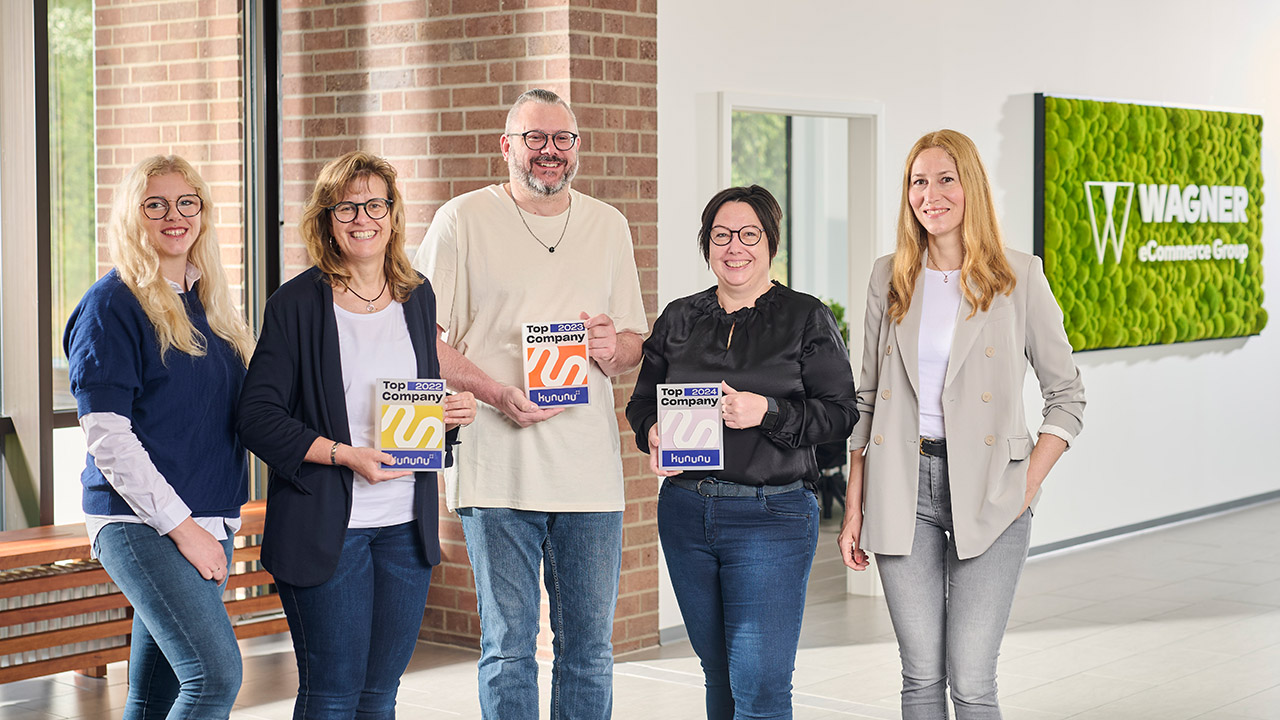 Fünf Menschen stehen in einem modernen Bürogebäude. Zwei von ihnen sind mit der Auszeichnung „Top Company“ ausgezeichnet. An der Wand hängt ein grünes Schild mit der Aufschrift „Wagner Group“. Sie lächeln und posieren neben großen Fenstern und einem grünen Logoschild.