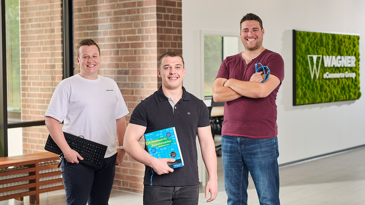 Das Bild zeigt drei Männer, die in einem modernen Bürogebäude vor einer Wand mit einem grünen Schild stehen, auf dem 'Wagner eCommerce Group' zu lesen ist. Der Mann links trägt ein weißes T-Shirt und hält eine Tastatur in der Hand. Der Mann in der Mitte trägt ein schwarzes Poloshirt und hält ein Buch mit dem Titel 'IT-Handbuch für Fachinformatiker'. Der Mann rechts trägt ein dunkelrotes T-Shirt und hält mehrere Kabel in der Hand. Alle drei Männer lächeln in die Kamera.