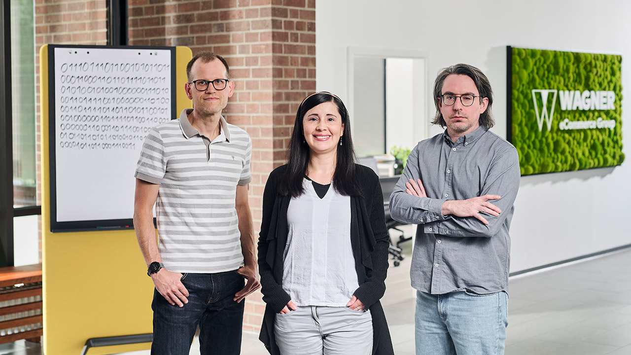 Das Bild zeigt drei Personen, die in einem modernen Bürogebäude vor einer Wand mit einem grünen Schild stehen, auf dem 'Wagner eCommerce Group' zu lesen ist. Links steht ein Mann mit Brille und einem grau-weiß gestreiften Poloshirt, der seine Hände in den Hosentaschen hat. In der Mitte steht eine Frau mit langen dunklen Haaren, einem weißen Oberteil und einer schwarzen Strickjacke, die in die Kamera lächelt. Rechts steht ein Mann mit Brille und schulterlangem Haar, der ein graues Hemd trägt und die Arme verschränkt hat. Im Hintergrund ist ein Whiteboard mit Binärcode-Muster zu sehen.