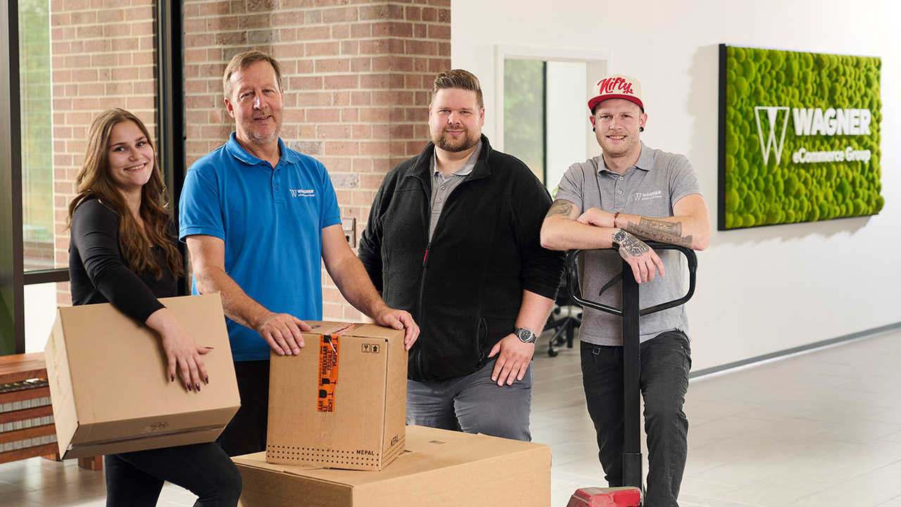 Das Bild zeigt vier Personen, die in einem modernen Bürogebäude vor einer Wand mit einem grünen Schild stehen, auf dem 'Wagner eCommerce Group' zu lesen ist. Links steht eine junge Frau mit langen braunen Haaren, die ein schwarzes Oberteil trägt und einen Karton in den Händen hält. Neben ihr steht ein älterer Mann in einem blauen Polo-Shirt mit dem Wagner-Logo, der ebenfalls einen Karton auf einem Tisch vor sich hält. Rechts von ihm steht ein Mann mit kurzem Haar und Bart, der eine schwarze Jacke trägt. Ganz rechts steht ein Mann in einem grauen Polo-Shirt mit dem Wagner-Logo und einer roten Baseballkappe, der einen Palettenhubwagen hält. Alle Personen lächeln in die Kamera.
