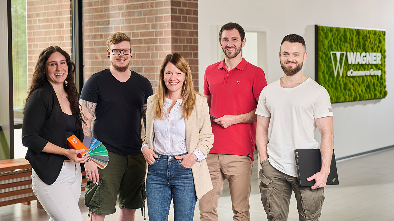 Das Bild zeigt fünf Personen, die in einem modernen Bürogebäude vor einer Wand mit einem grünen Schild stehen, auf dem 'Wagner eCommerce Group' zu lesen ist. Links steht eine Frau mit lockigem braunem Haar, die ein schwarzes Oberteil und eine weiße Hose trägt. Sie hält Farbpaletten in der Hand. Neben ihr steht ein Mann mit Brille und kurzem Haar, der ein schwarzes T-Shirt und olivgrüne Shorts trägt. In der Mitte steht eine Frau mit langen braunen Haaren, die ein beiges Sakko, ein weißes Hemd und eine blaue Jeans trägt. Rechts von ihr steht ein Mann mit einem roten Poloshirt, der ein Tablet in der Hand hält. Ganz rechts steht ein Mann mit kurzem Haar und Bart, der ein weißes T-Shirt und khakifarbene Hosen trägt und einen Laptop in der Hand hält. Alle Personen lächeln in die Kamera