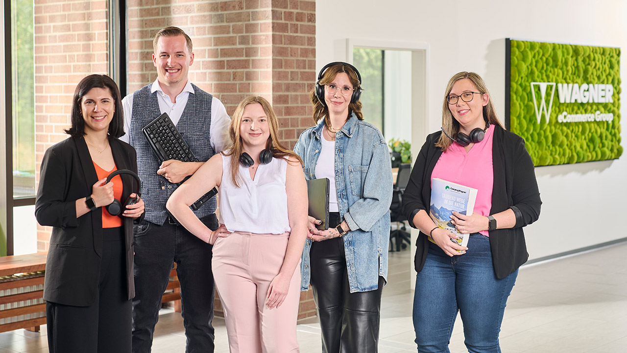 Fünf lächelnde Personen in einem Büro, halten Kopfhörer, Tastatur, Laptop und Magazin, vor einem Wagner eCommerce Schild.