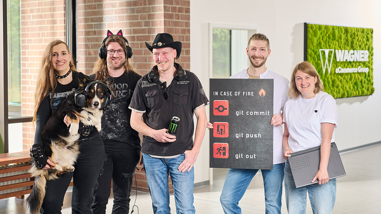 Das Bild zeigt sechs Personen, die in einem modernen Bürogebäude vor einer Wand mit einem grünen Schild stehen, auf dem 'Wagner eCommerce Group' zu lesen ist. Links steht eine Frau mit langen braunen Haaren, die ein schwarzes Metal-T-Shirt trägt und einen Hund hält, der Kopfhörer trägt. Neben ihr steht ein Mann mit langen Haaren und einer Katzenohren-Kopfbedeckung, der ebenfalls ein schwarzes T-Shirt trägt. Daneben steht ein Mann mit Cowboyhut und Kopfhörern um den Hals, der eine Dose Monster-Energy in der Hand hält. Rechts von ihm steht ein Mann, der ein Schild mit der Aufschrift 'In Case of Fire: git commit, git push, git out' hält. Ganz rechts steht eine Frau mit blonden Haaren und weißem T-Shirt, die ein Laptop unter dem Arm hält. Alle Personen lächeln in die Kamera.