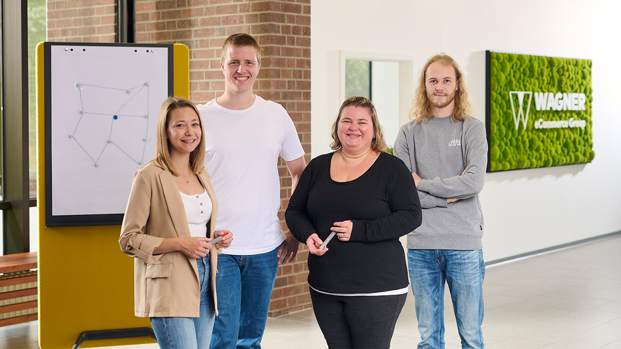 Eine Gruppe von vier lächelnden Personen steht in einem modernen Büro. Sie befinden sich vor einem Whiteboard mit einer einfachen geometrischen Zeichnung und einem grünen Wandschild, auf dem 'Wagner eCommerce Group' steht.