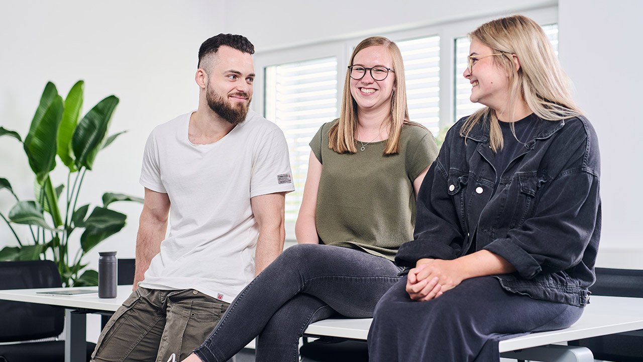 Drei lächelnde Personen sitzen auf einem Tisch in einem hellen Raum, sie unterhalten sich entspannt miteinander.