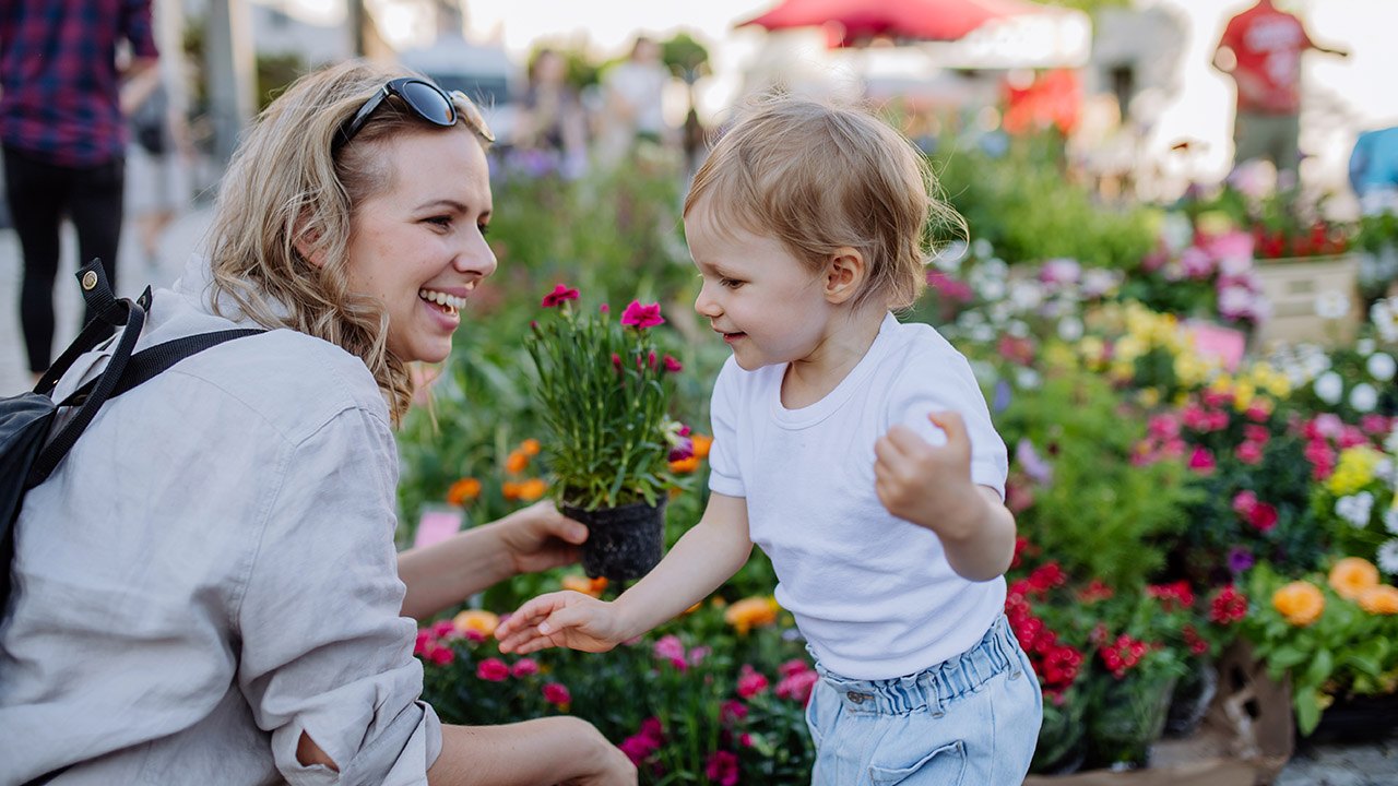Lächelnde Frau zeigt einem kleinen Kind Blumen auf einem bunten Markt, beide wirken glücklich und entspannt.