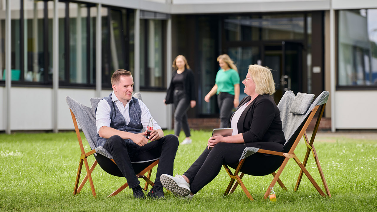 Zwei Personen sitzen entspannt in Liegestühlen auf einer Wiese vor einem Bürogebäude und unterhalten sich, während zwei weitere Personen im Hintergrund vorbeigehen.