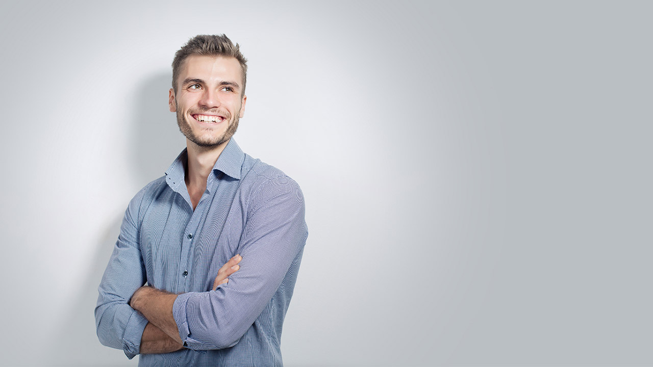 Lächelnder Mann in einem blauen Hemd steht mit verschränkten Armen vor einem hellen Hintergrund, blickt zur Seite.