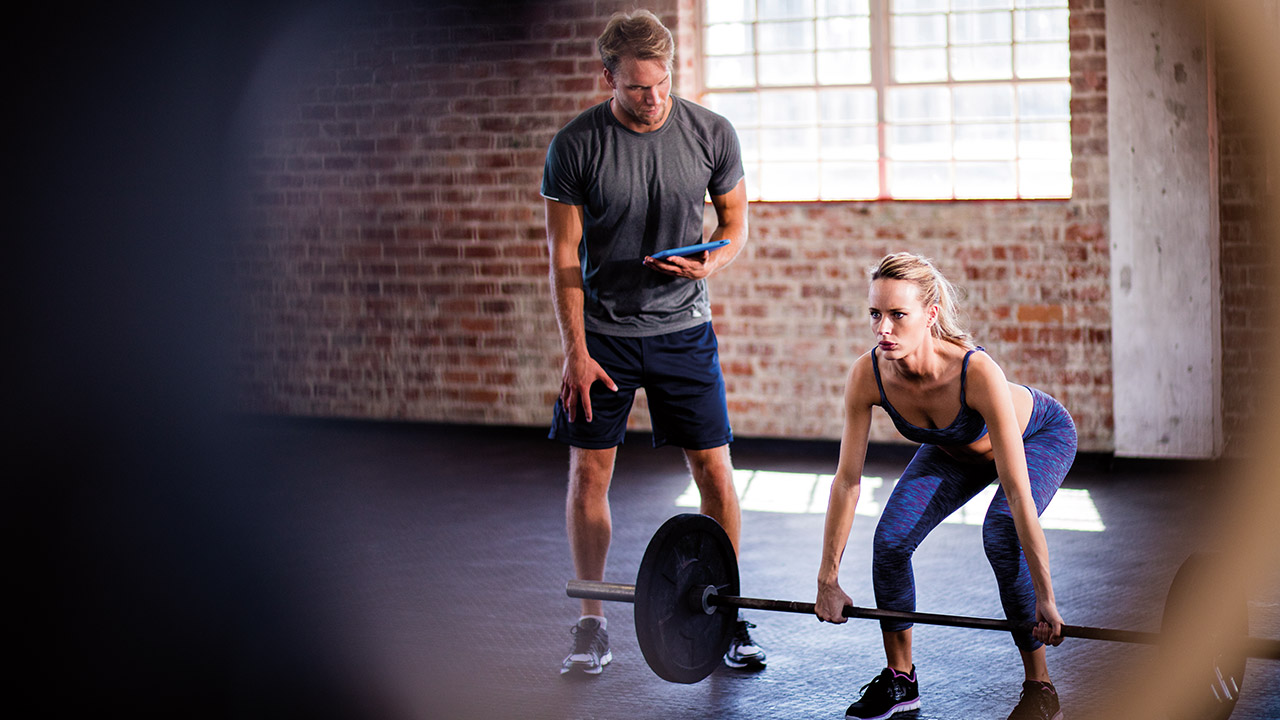 Trainer beobachtet eine Frau beim Gewichtheben in einem Fitnessstudio mit Backsteinwänden, beide konzentriert bei der Arbeit.