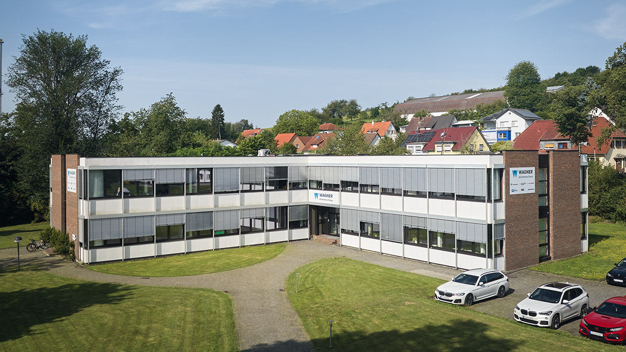 Außenansicht eines modernen Bürogebäudes der Wagner eCommerce Group, umgeben von Rasenflächen und parkenden Autos.