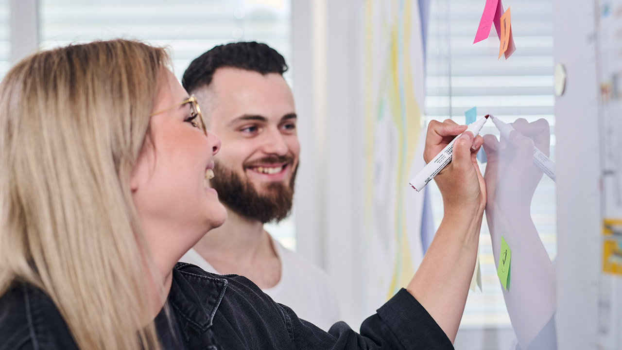 Zwei Personen lächeln, während sie gemeinsam an einer Glaswand mit Stift und Haftnotizen arbeiten.