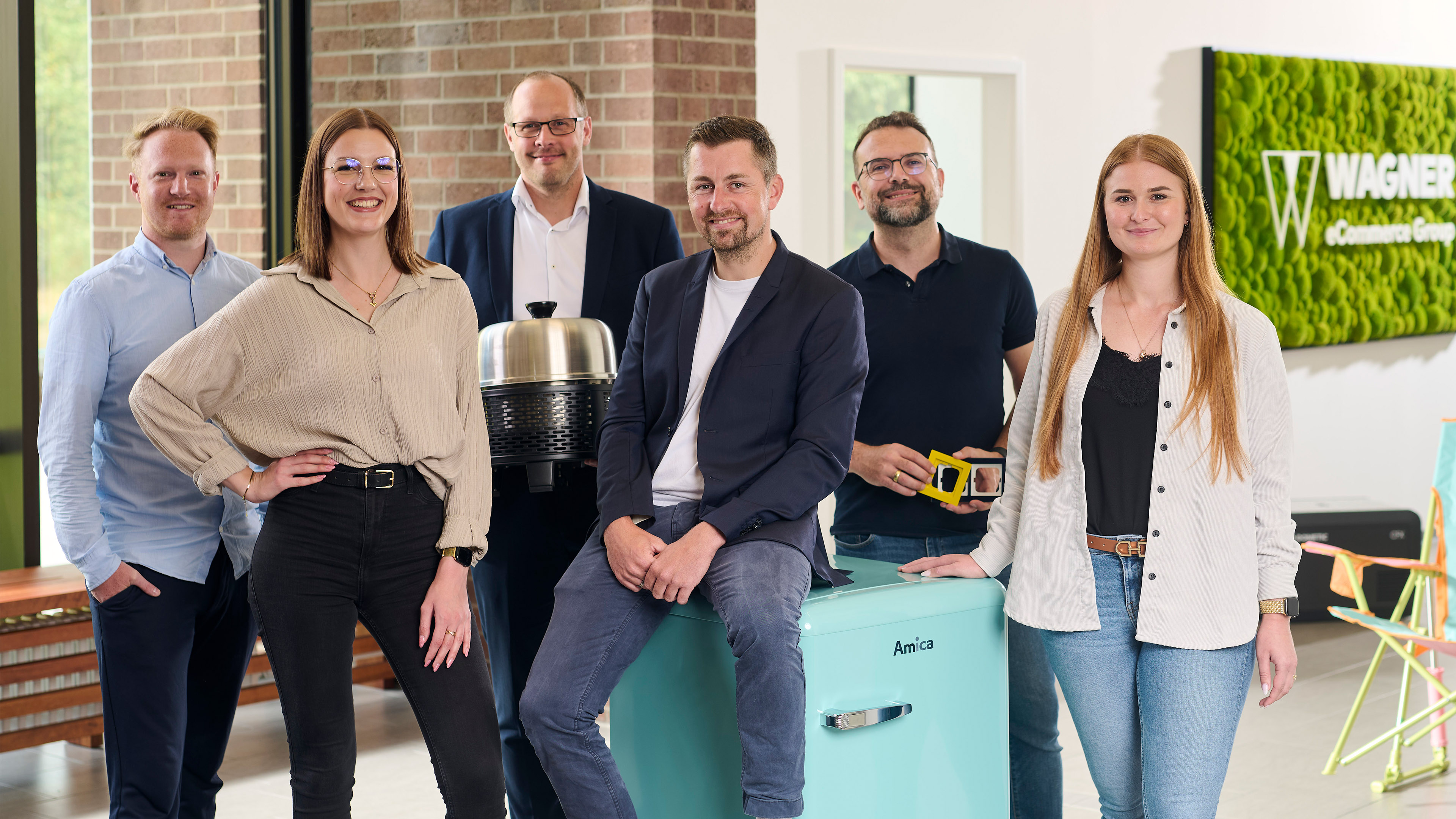 Fünf Personen stehen in einem Büro und halten jeweils eine Nummernkarte von 1 bis 4 in der Hand. Im Hintergrund ist ein Whiteboard mit Zahlen zu sehen. Das Büro ist modern gestaltet und hat eine grüne Pflanzenwand.