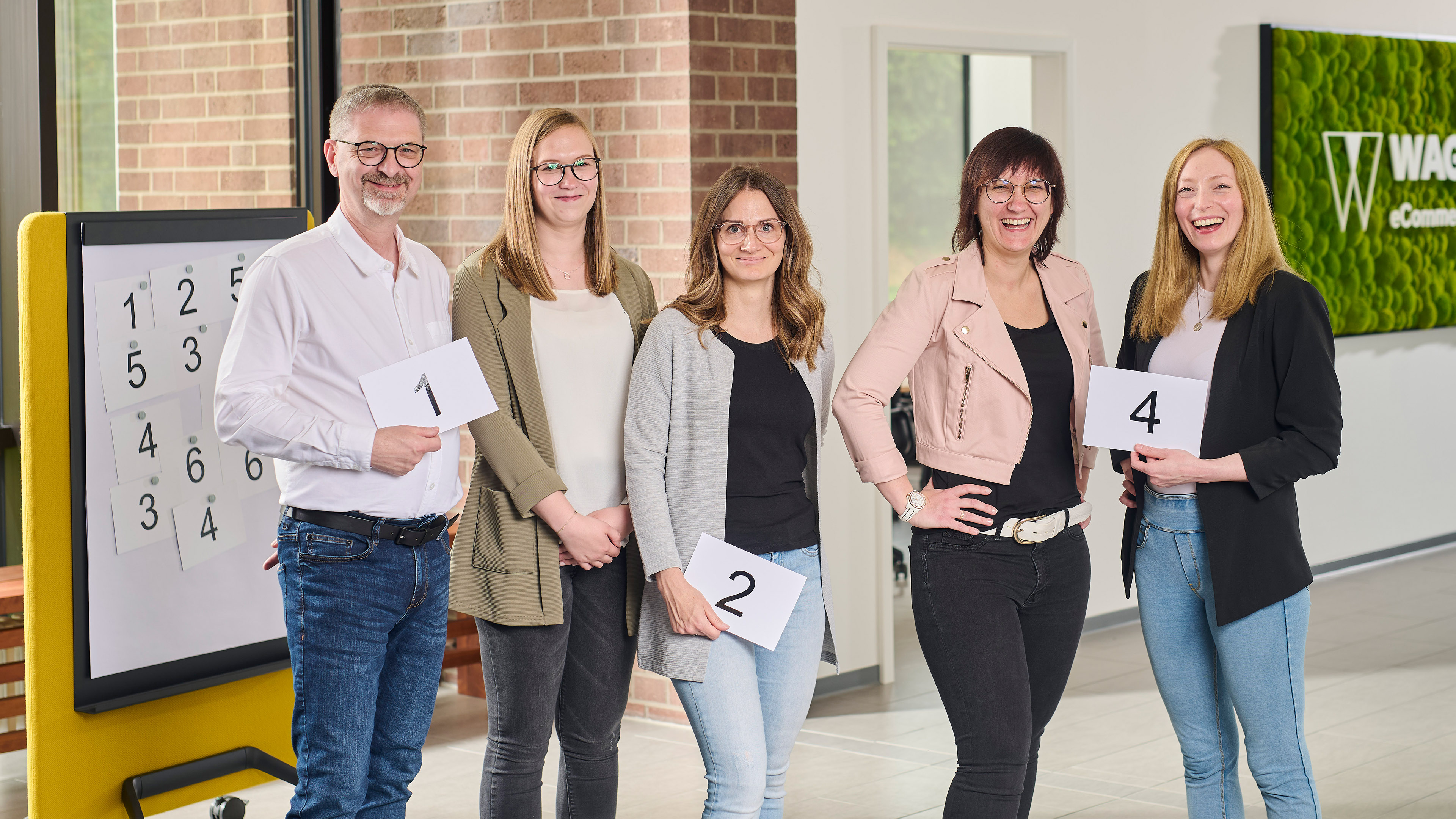 Ein Team von fünf Personen posiert mit nummerierten Karten in einem hellen Büro. Ein Zahlenbrett und ein grünes Wandschild mit der Aufschrift 'Wagner eCommerce Group' sind im Hintergrund sichtbar.