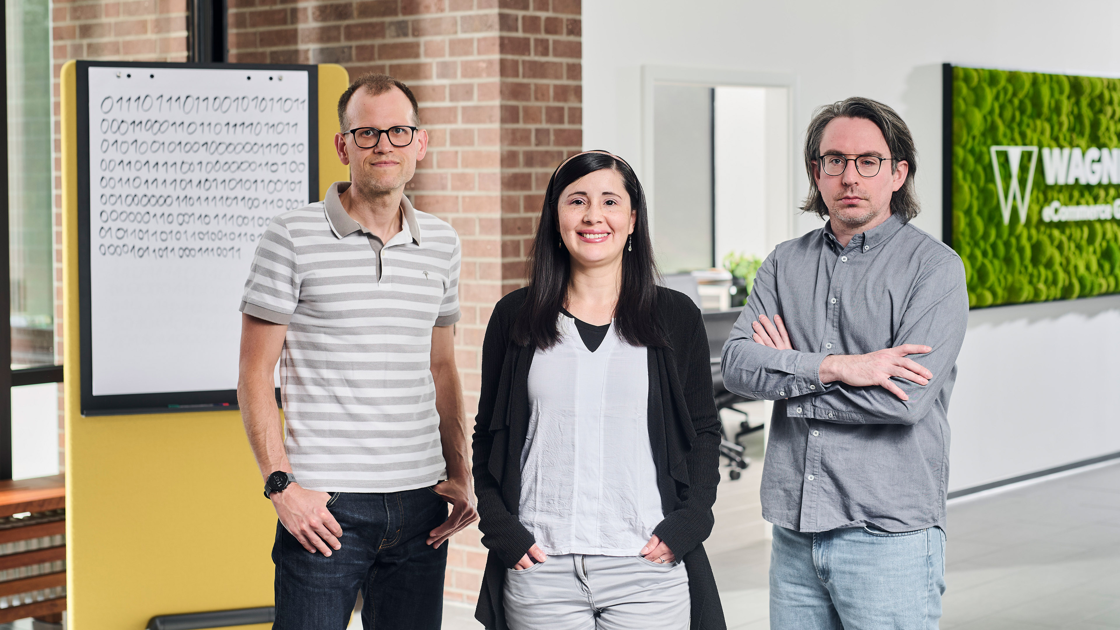 Das Bild zeigt drei Personen, die in einem modernen Bürogebäude vor einer Wand mit einem grünen Schild stehen, auf dem 'Wagner eCommerce Group' zu lesen ist. Links steht ein Mann mit Brille und einem grau-weiß gestreiften Poloshirt, der seine Hände in den Hosentaschen hat. In der Mitte steht eine Frau mit langen dunklen Haaren, einem weißen Oberteil und einer schwarzen Strickjacke, die in die Kamera lächelt. Rechts steht ein Mann mit Brille und schulterlangem Haar, der ein graues Hemd trägt und die Arme verschränkt hat. Im Hintergrund ist ein Whiteboard mit Binärcode-Muster zu sehen.